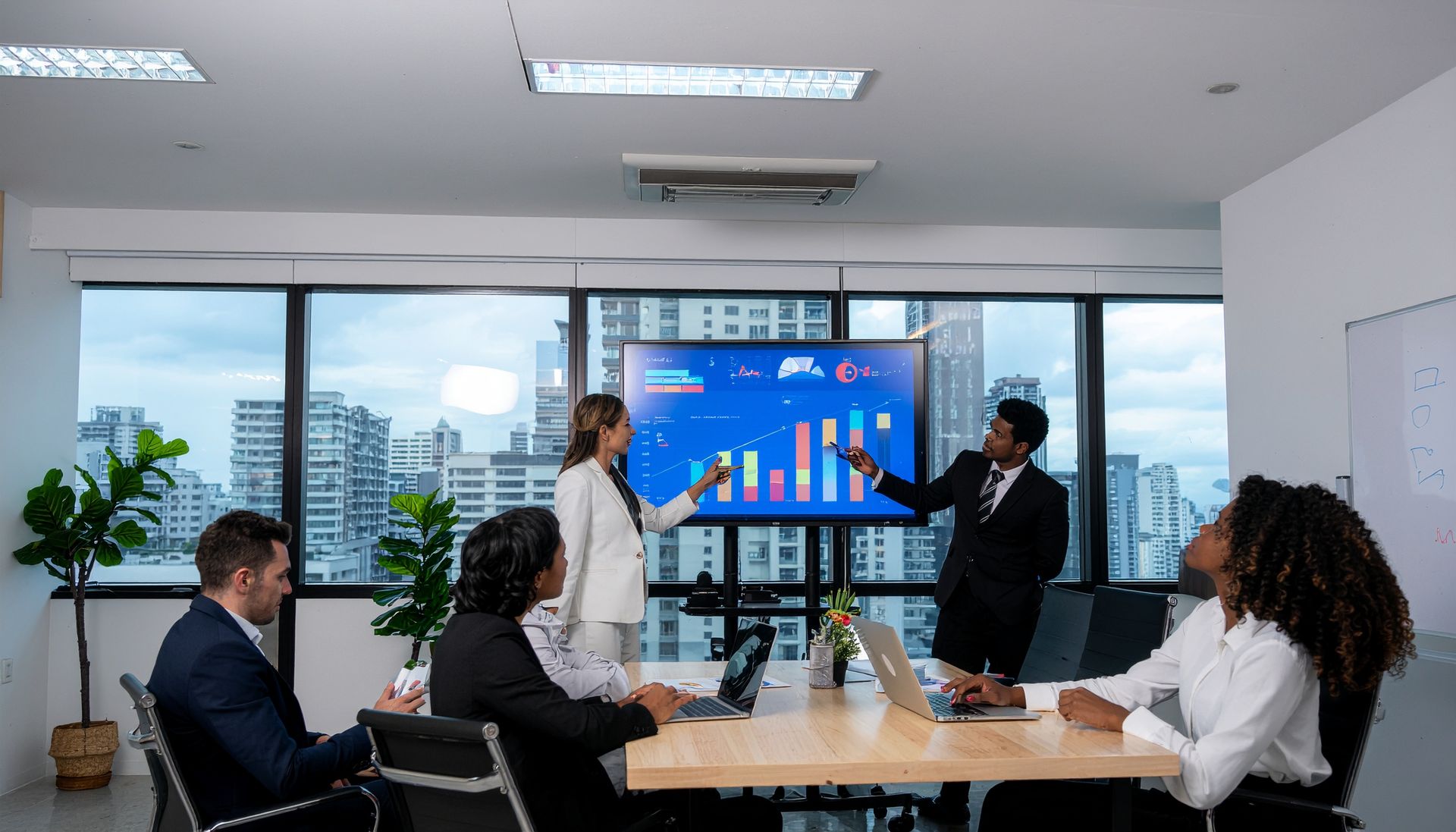 Business meeting in a bright conference room, with people presenting charts on a large screen.