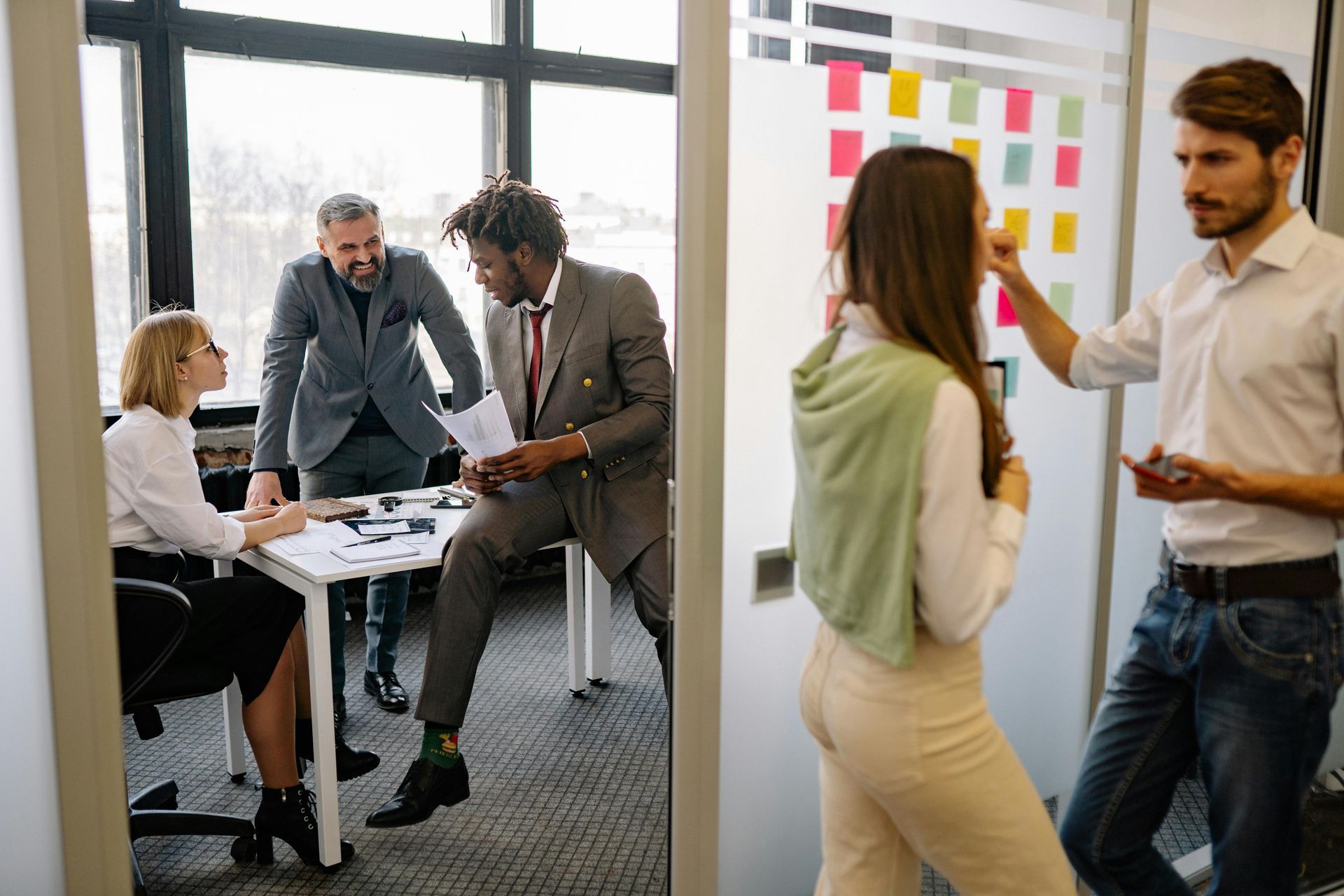 A professional group collaborates in a bright office, with some members working at a table and others chatting nearby.