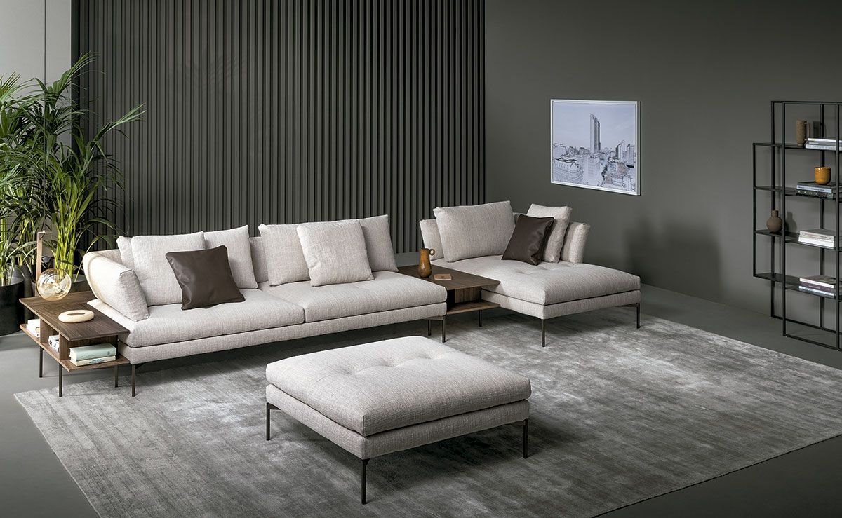 Sala de estar moderna con sofá seccional beige, otomana, alfombra gris y estanterías sobre paredes oscuras.