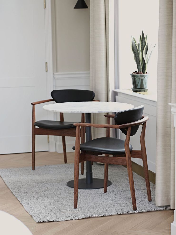 Pequeño rincón comedor con mesa redonda, sillas de madera, alfombra gris y una planta en maceta junto a la ventana.