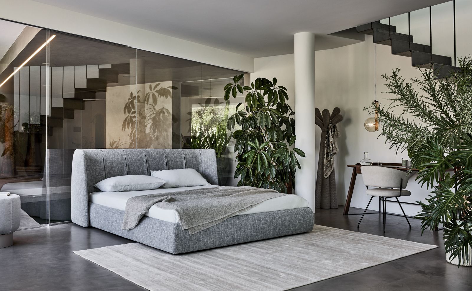 Modern bedroom with a low gray bed, striped rug, plants, and glass stairs in an open interior.
