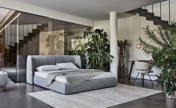 Dormitorio moderno con cama gris, escalera de cristal, plantas grandes y una elegante zona de escritorio.