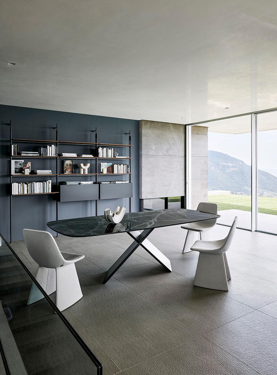 Comedor moderno con mesa escultural, sillas blancas, estantes oscuros y ventanales que van del suelo al techo