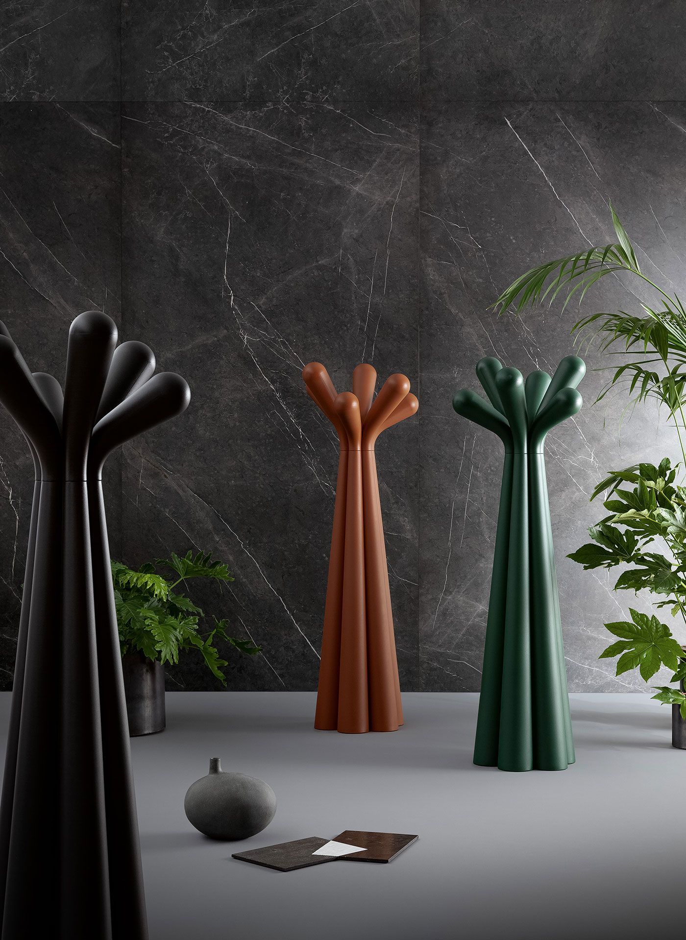 Tres percheros esculturales en negro, óxido y verde en una habitación moderna con plantas en macetas y una pared oscura 