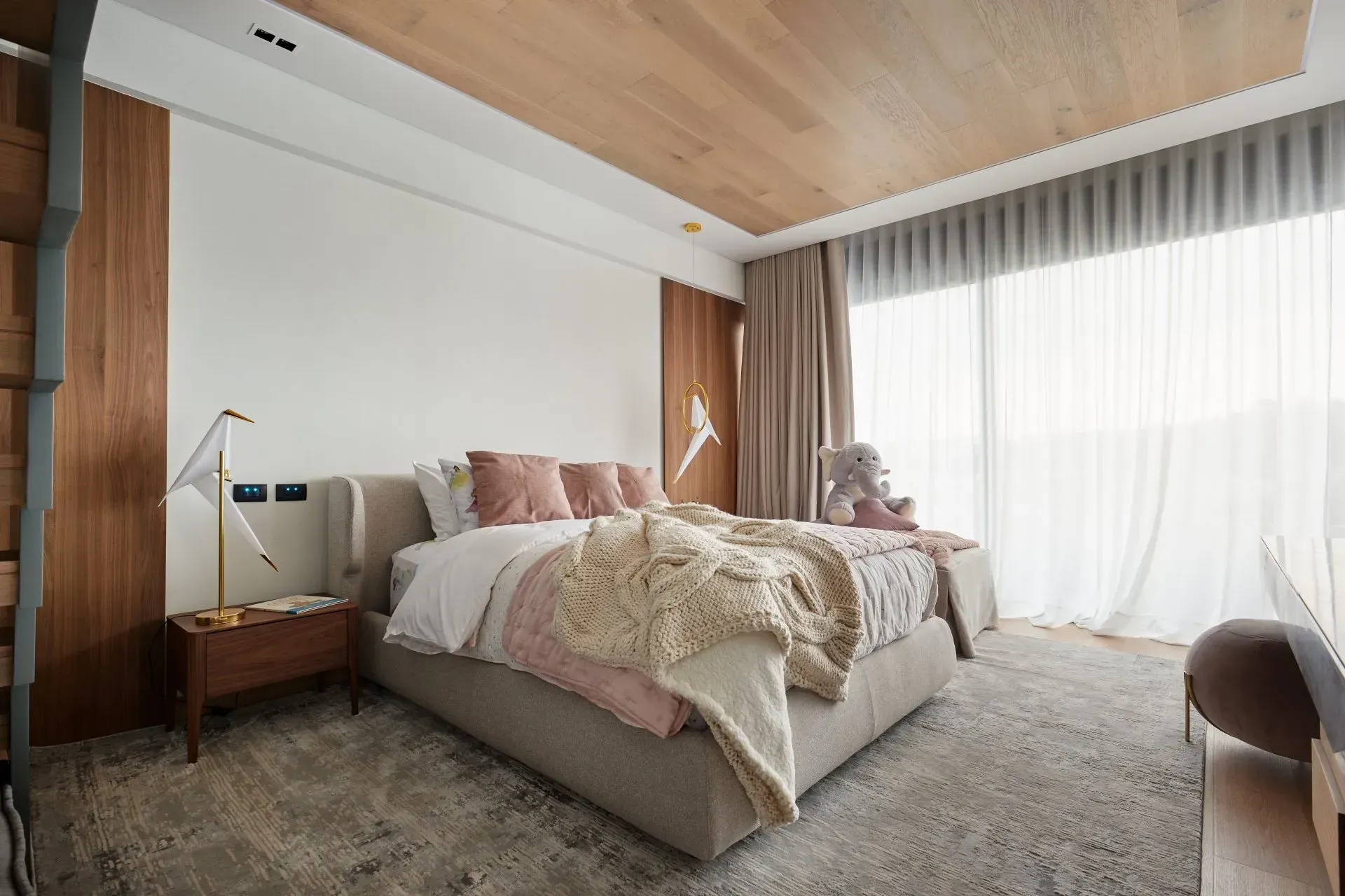 Dormitorio moderno con una cama grande, techo revestido de madera, decoración neutra y ventanales del suelo al techo.