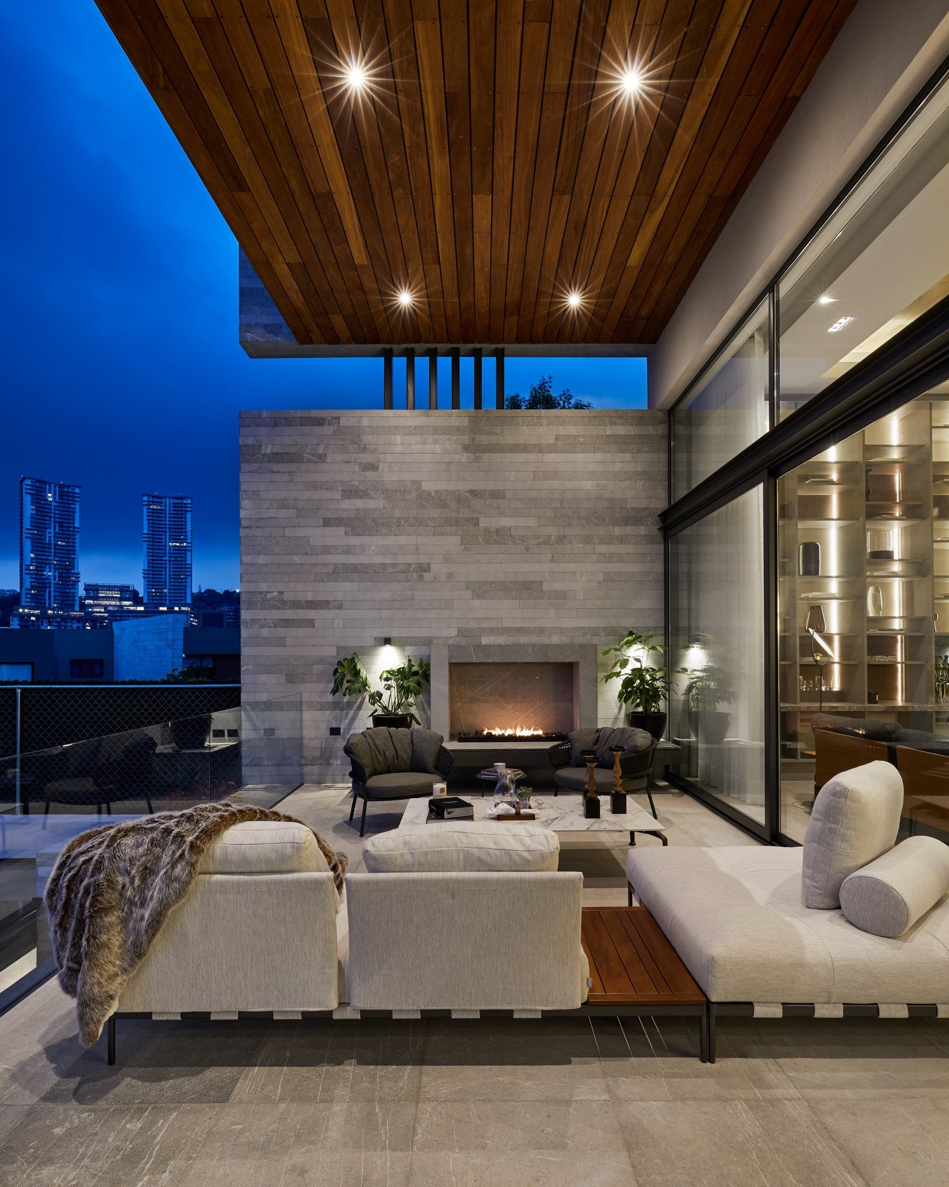 Patio moderno de lujo con asientos blancos, chimenea y vistas al horizonte de la ciudad al atardecer.