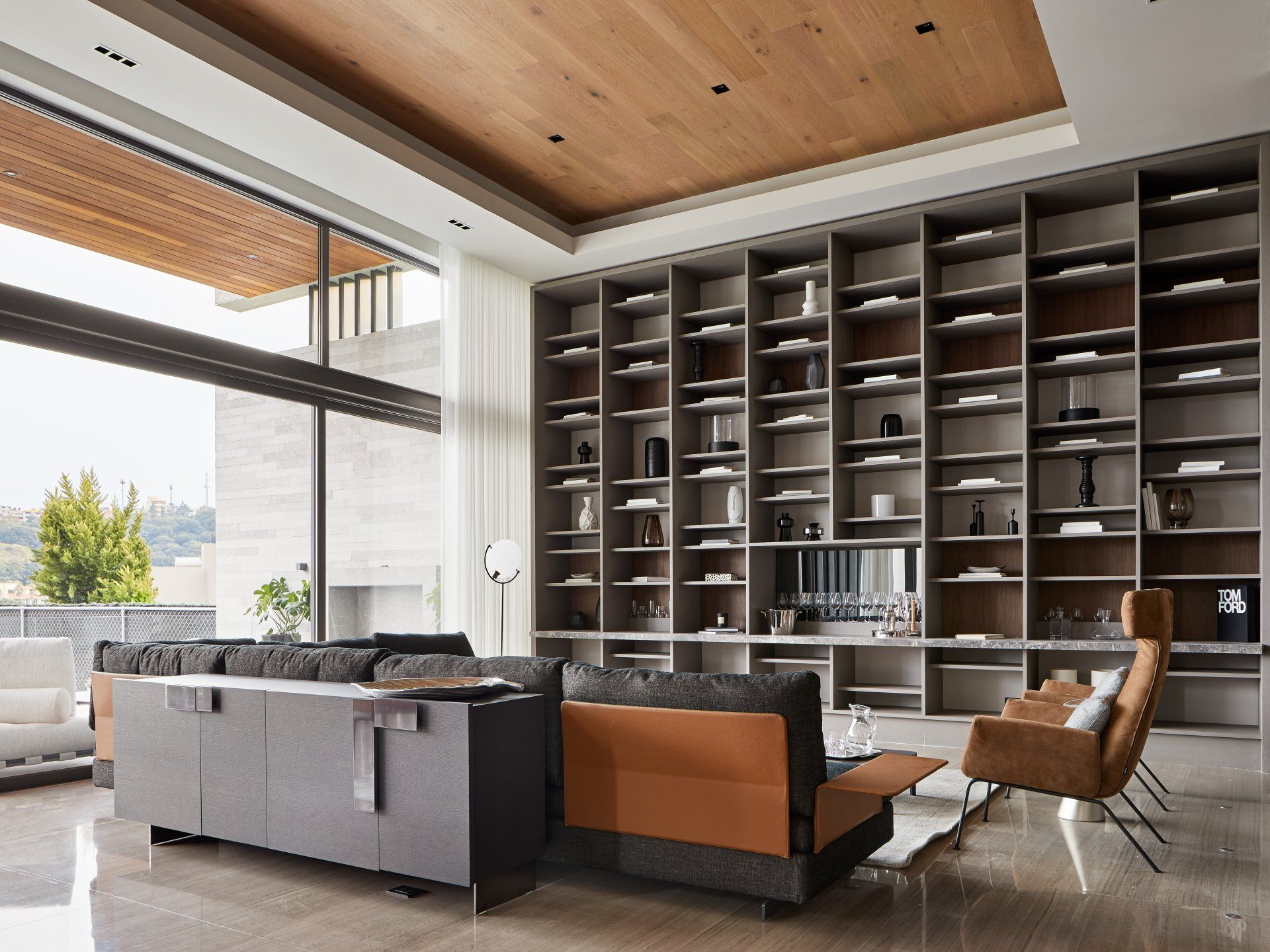 Sala de estar moderna con grandes estantes de pared, sofá gris, sillón color canela y ventanales del suelo al techo.