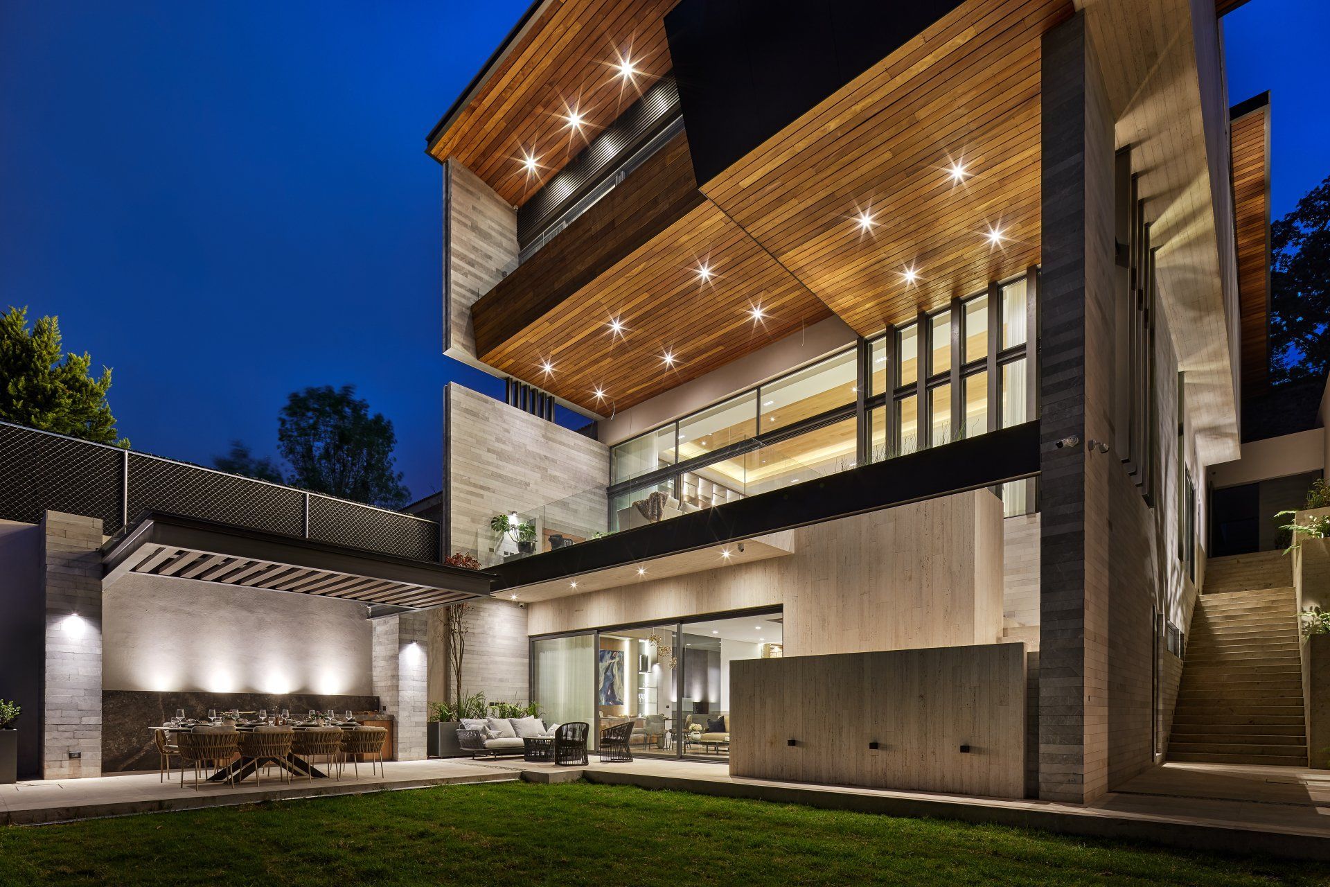 Casa moderna de dos plantas al atardecer con iluminación interior cálida, detalles en madera y un jardín paisajístico.
