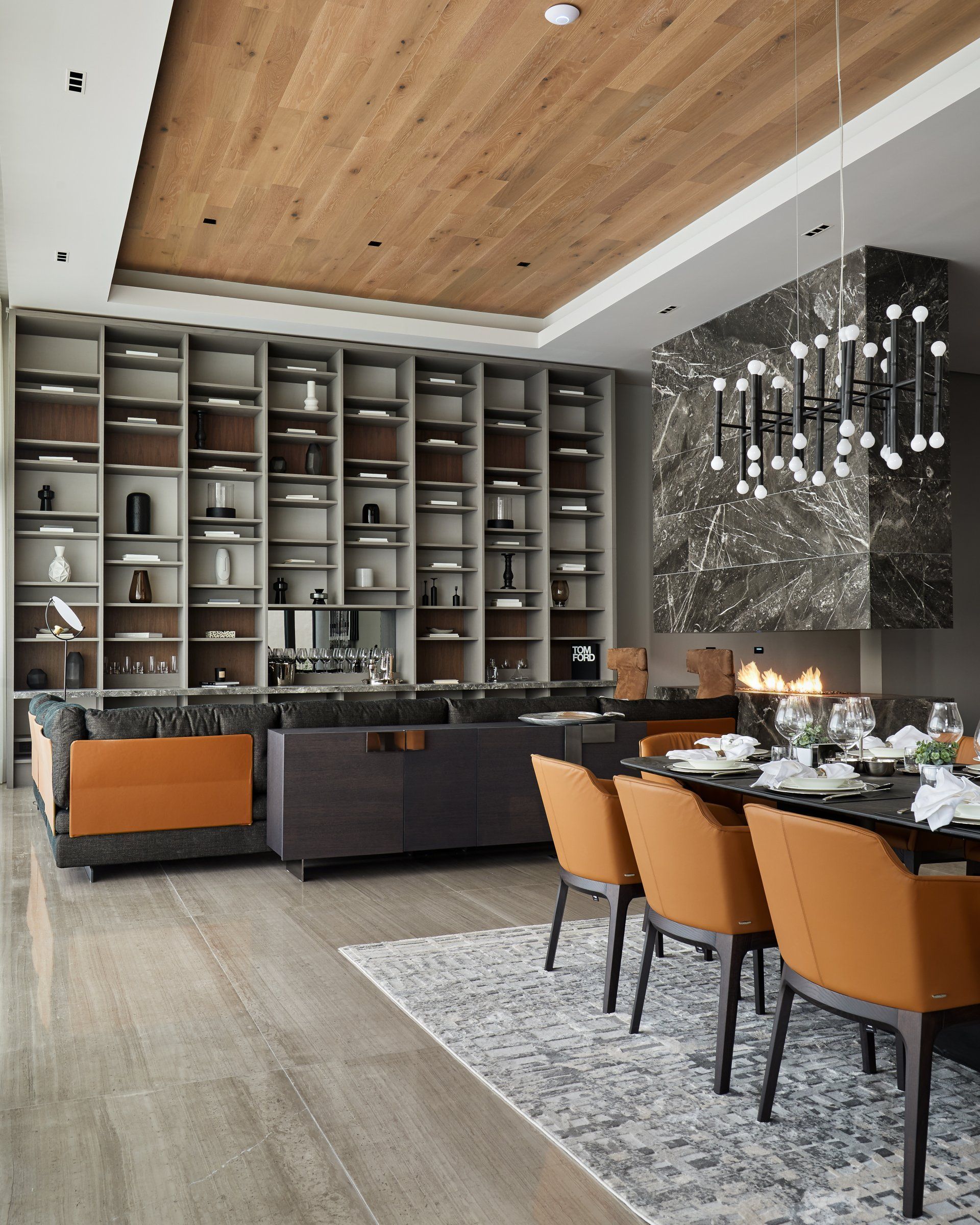 Comedor moderno con mesa larga, sillas naranjas, sofá oscuro, estanterías abiertas y una lámpara de araña
