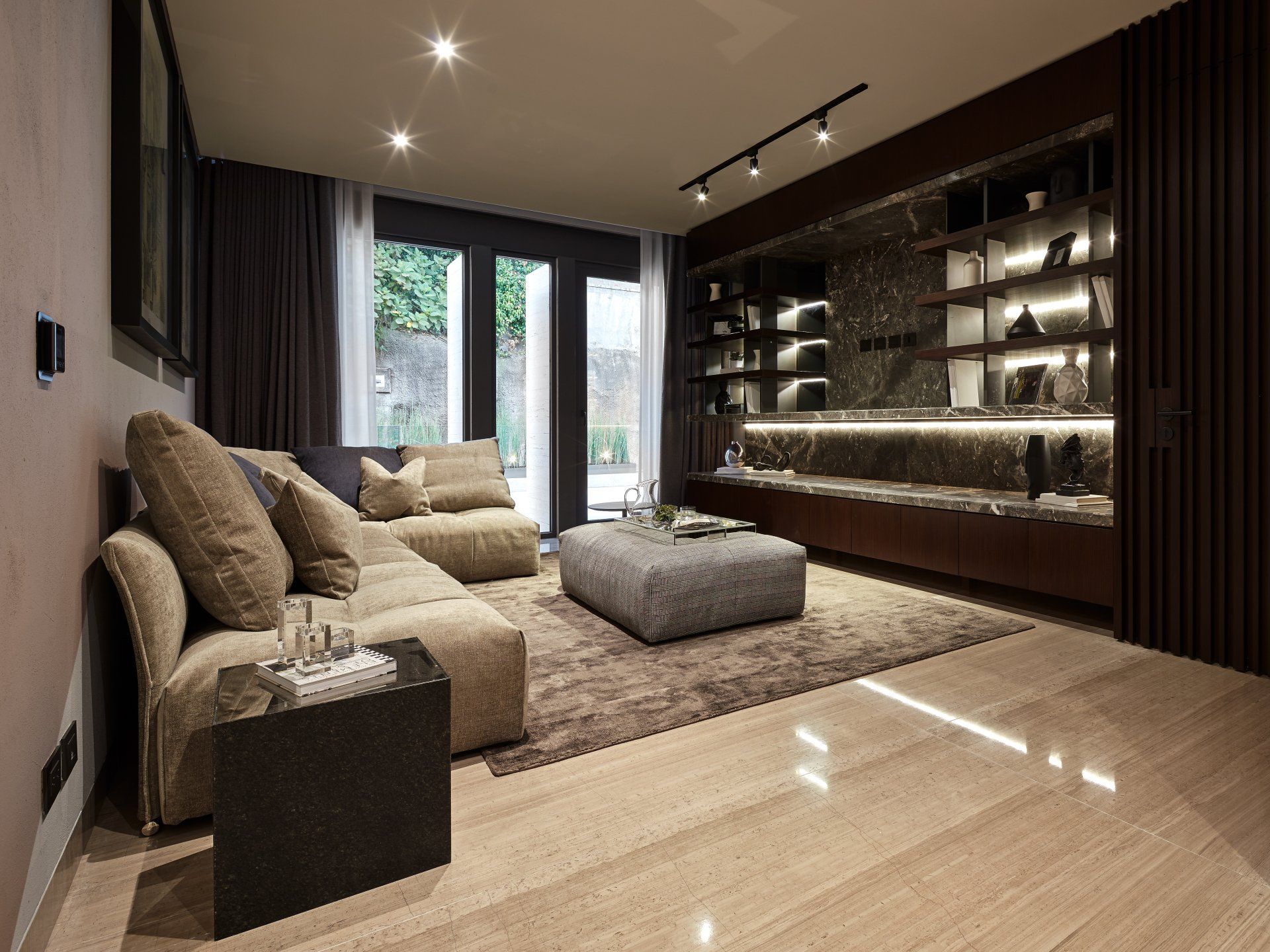 Sala de estar moderna con sofá beige, otomana, estanterías de madera oscura y grandes ventanales.