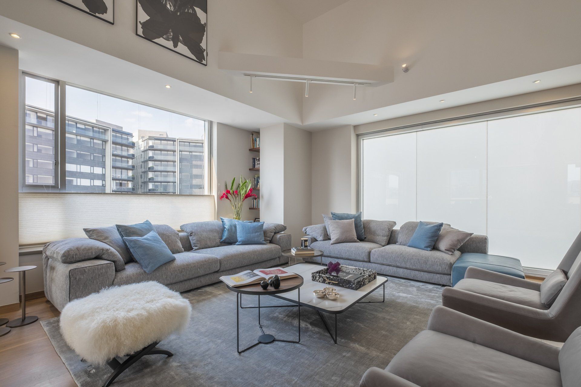Sala de estar moderna con sofá seccional gris, sillas blancas, mesa de centro y grandes ventanales con vistas a edificios