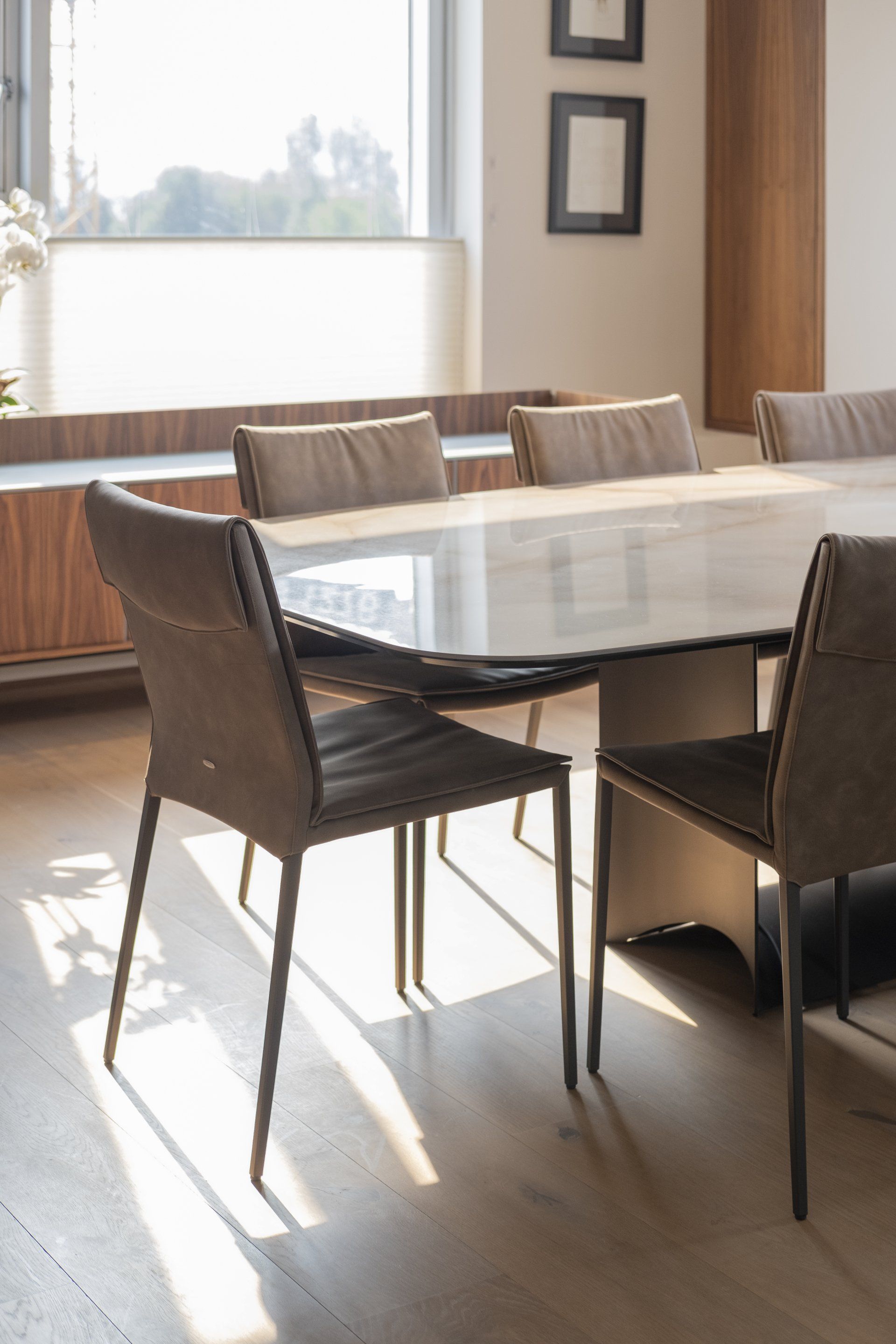 Comedor vacío con mesa de cristal, sillas de mimbre y luz solar que entra por una gran ventana.