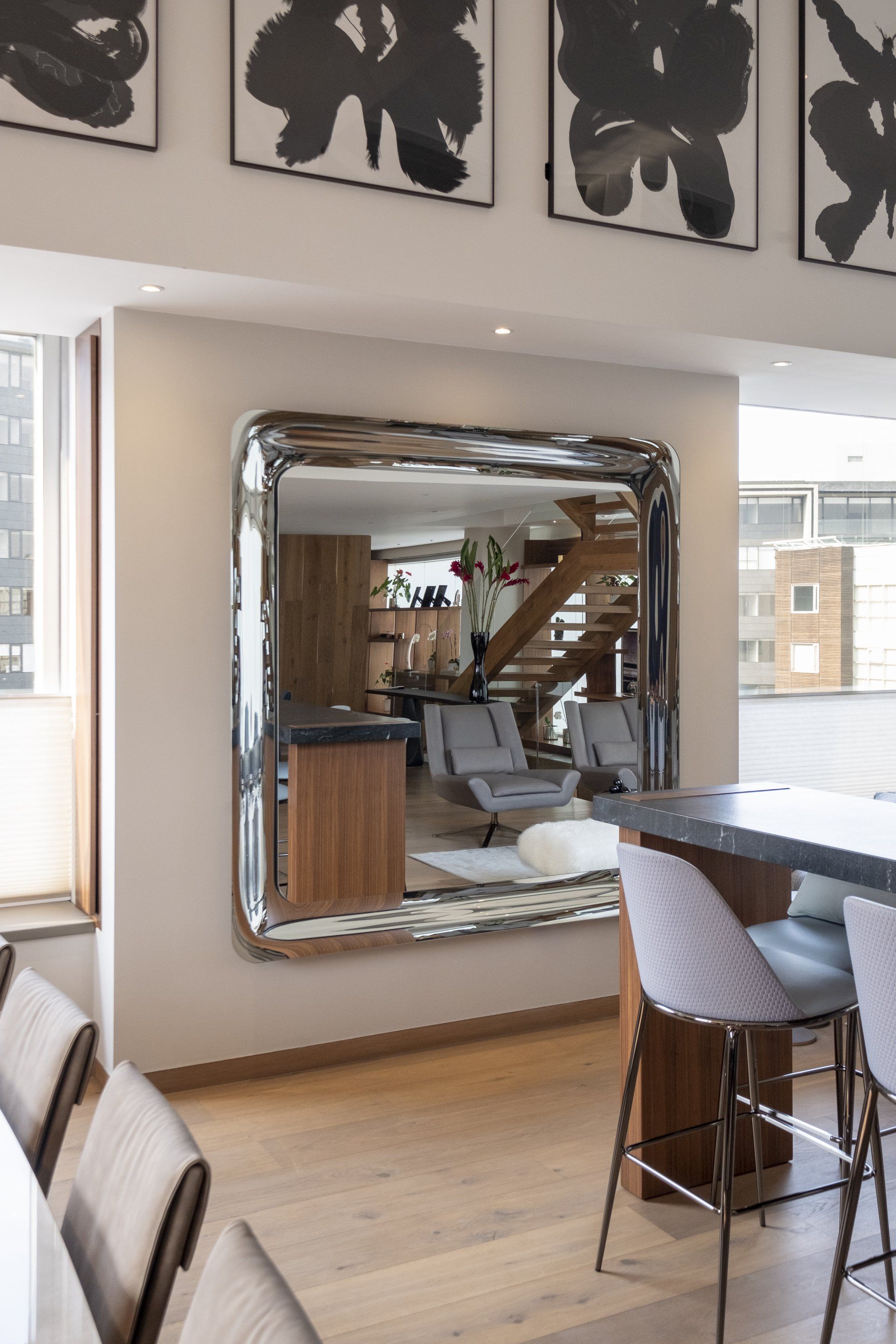 Comedor moderno con una gran pared decorada con espejos, suelo de madera y una mesa con taburetes junto a la ventana.