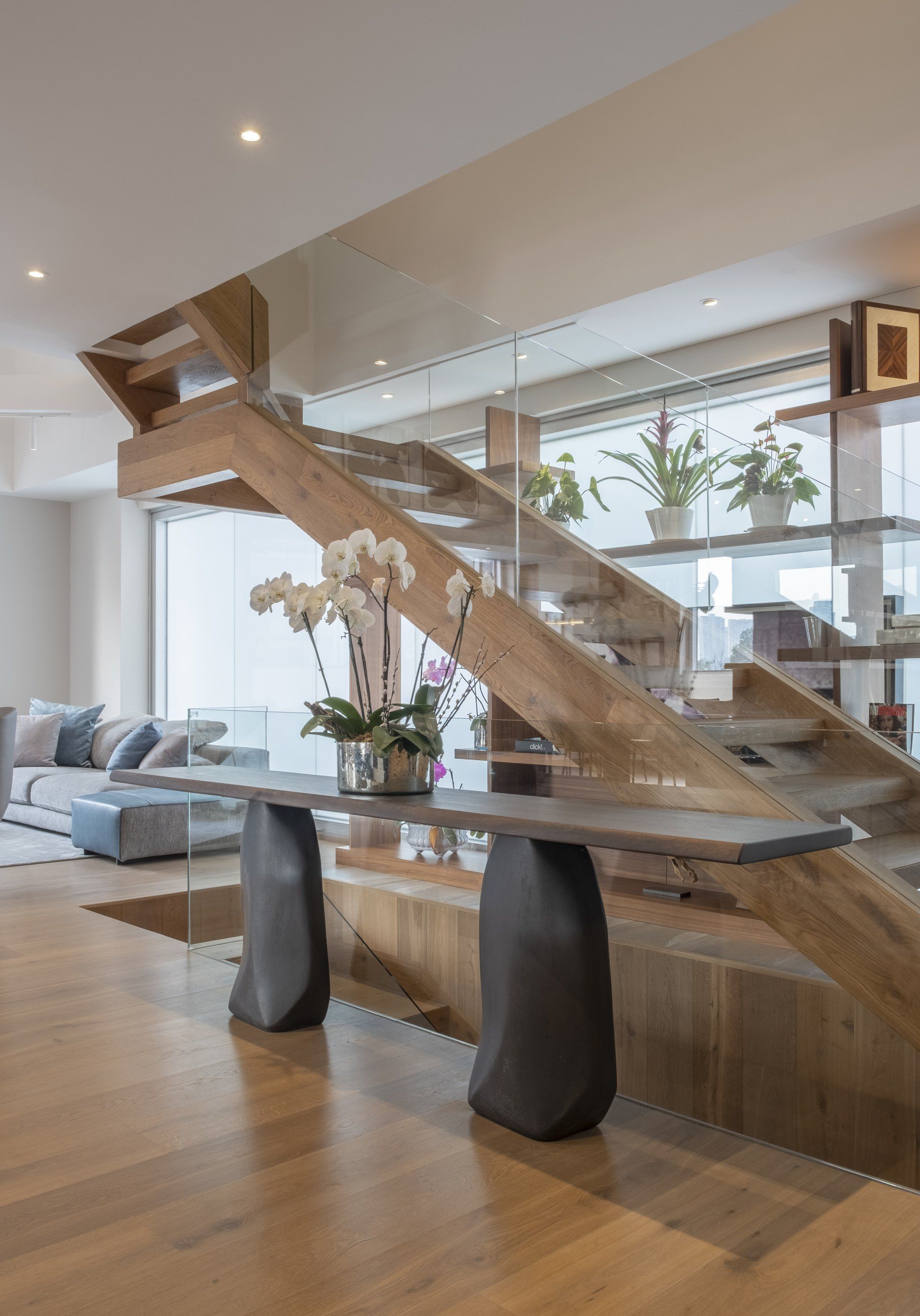 Sala de estar moderna con una escalera de madera escultural y un altillo con barandilla de cristal sobre un sofá y una mesa