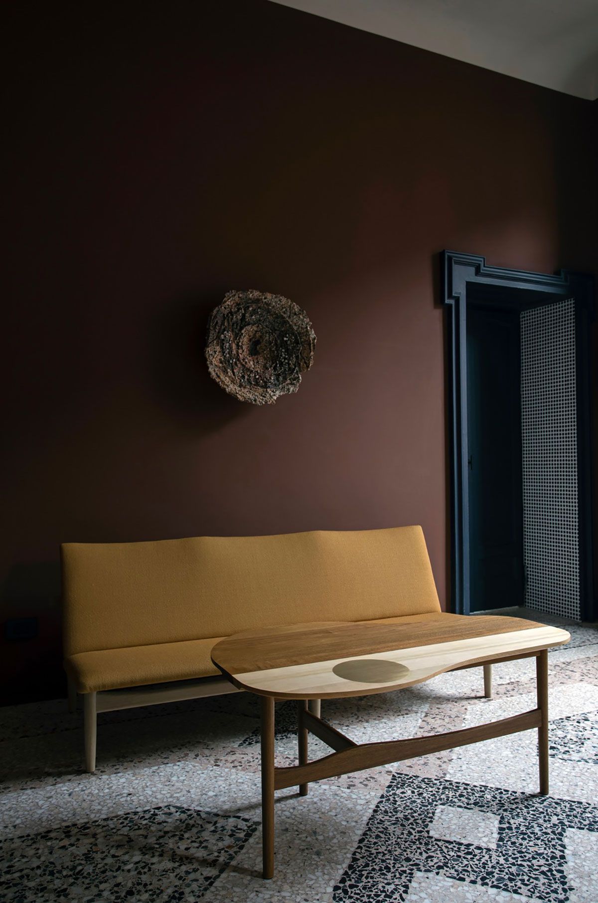 Patio moderno con banco color mostaza, mesa de centro redonda y pared oscura con decoración colgante.