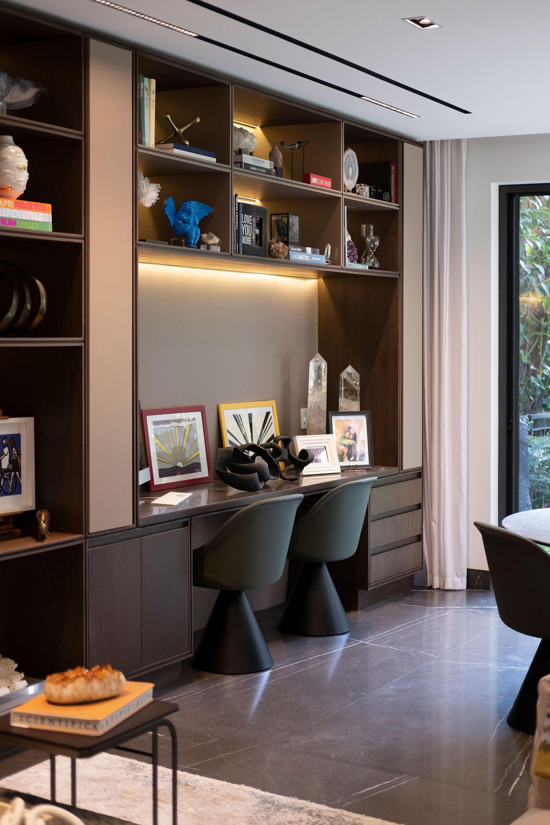 Rincón de oficina moderno en casa con estanterías empotradas, dos sillas y un escritorio junto a una gran ventana.
