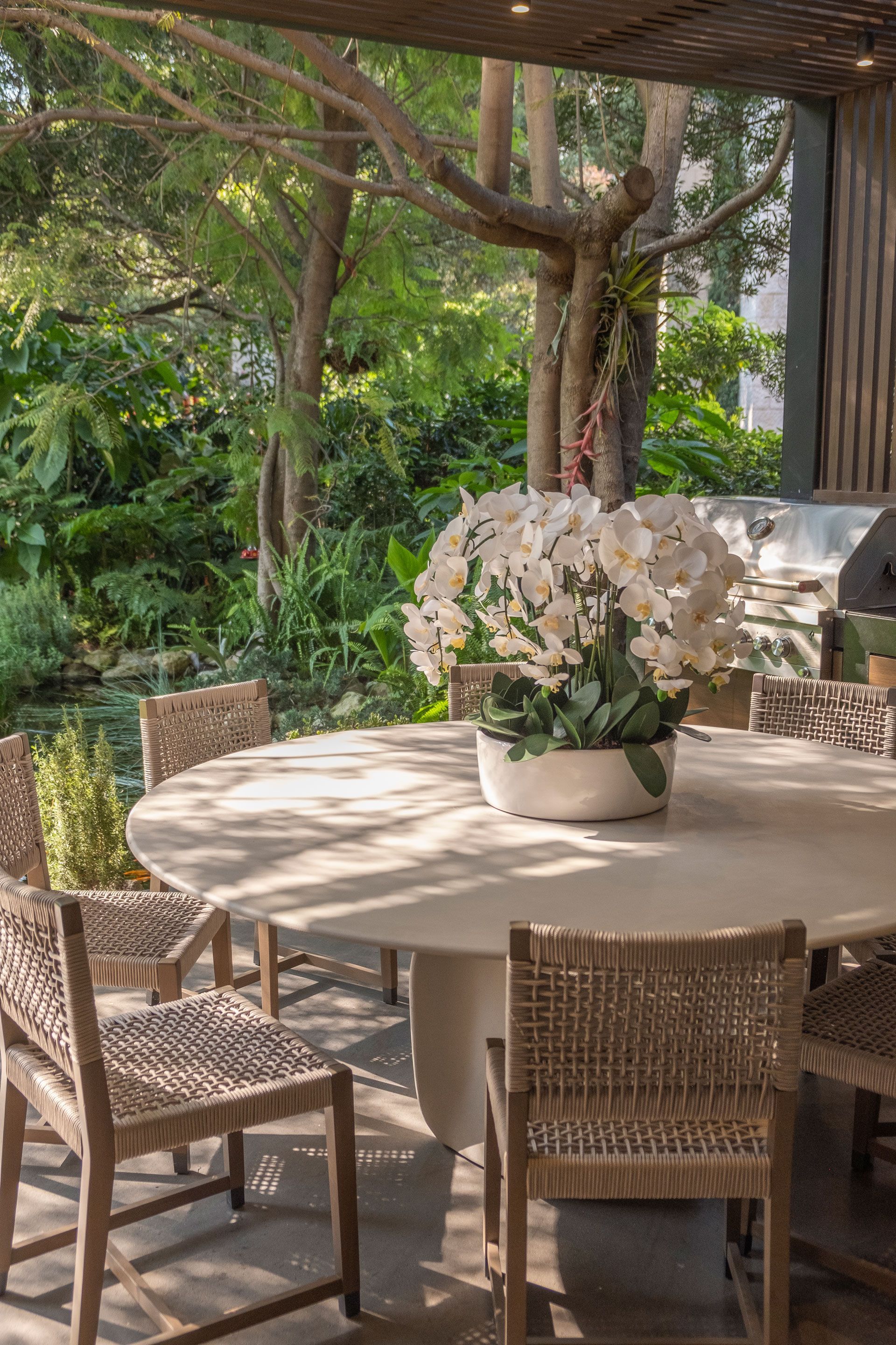 Mesa de comedor exterior para patio con sillas de mimbre y un centro de mesa floral blanco bajo una pérgola con sombra.
