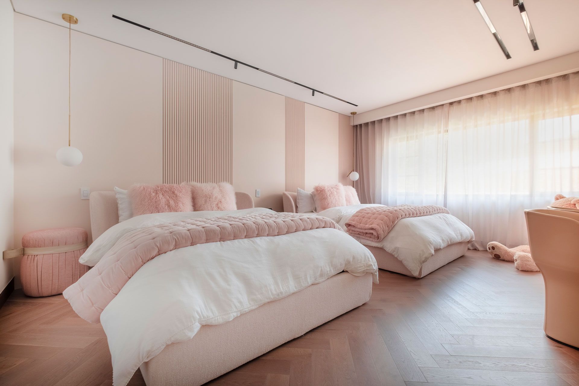 Habitación de color rosa pálido con dos camas sin hacer, cortinas transparentes y luz natural cálida.