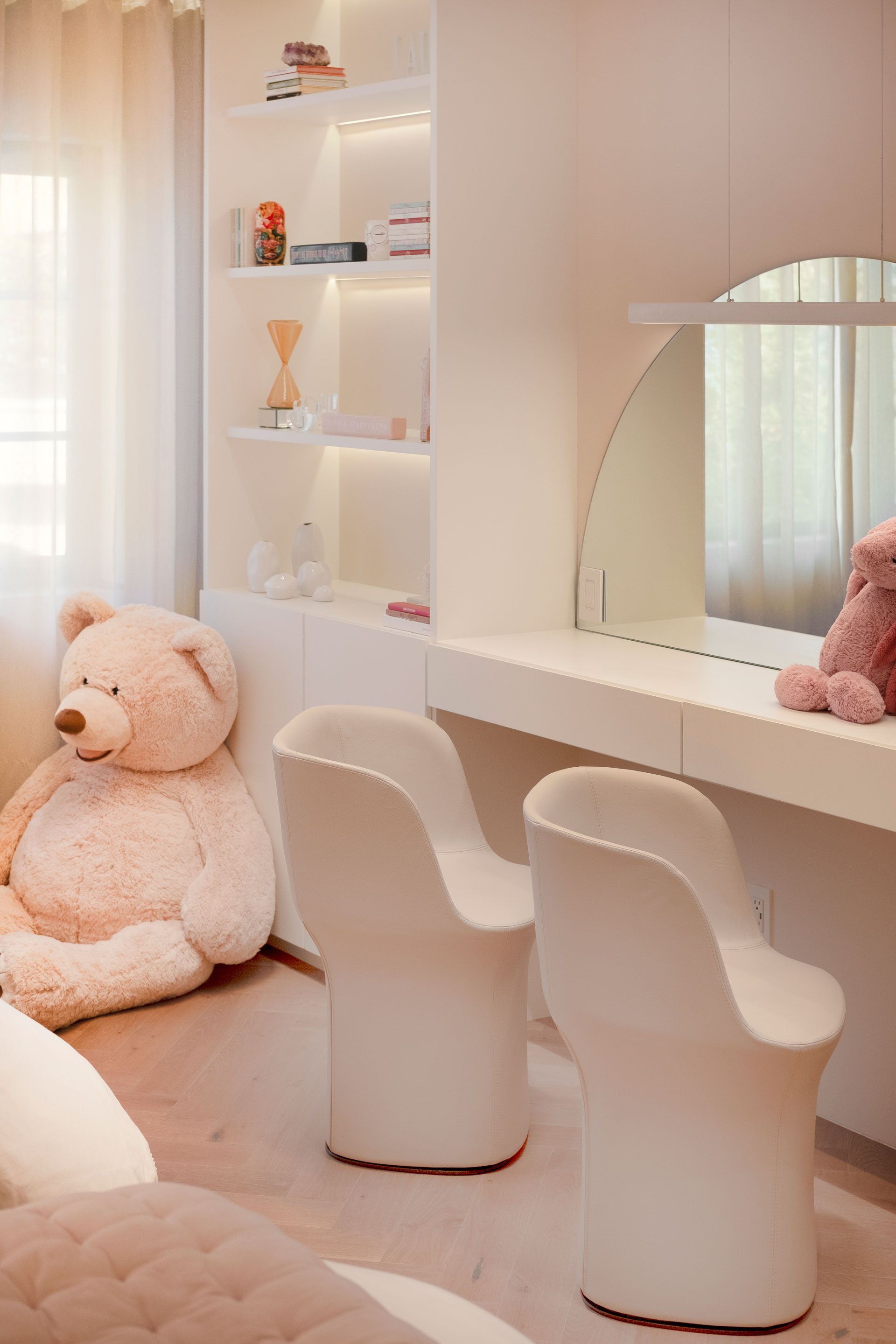 Tocador rosa con un gran oso de peluche, sillas blancas, estantes y un espejo redondo.