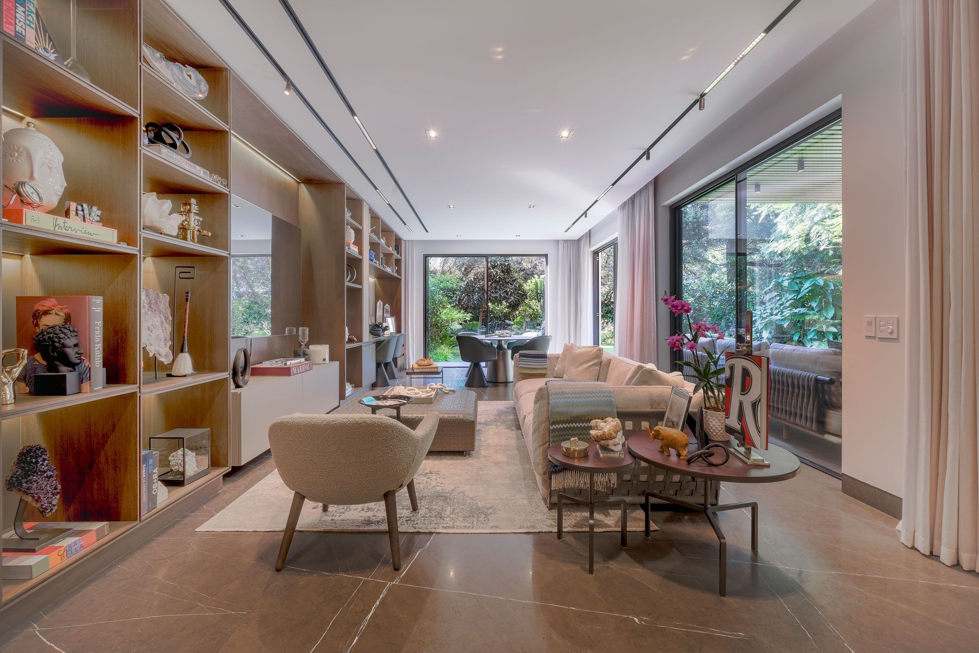 Salón moderno con asientos beige, estanterías, grandes ventanales y una mesa de centro con vistas al jardín.