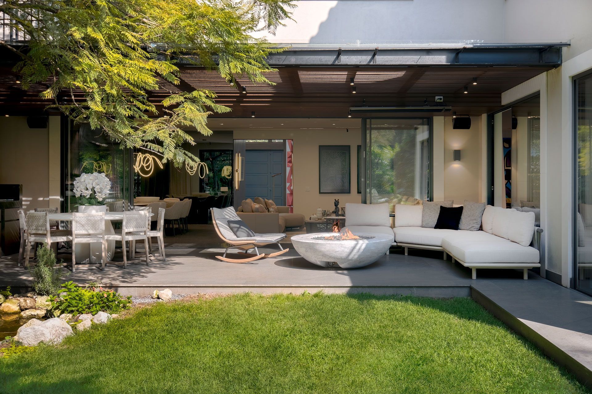 Patio moderno con sofá blanco de exterior, tumbonas, juego de comedor y césped verde bajo una terraza cubierta.