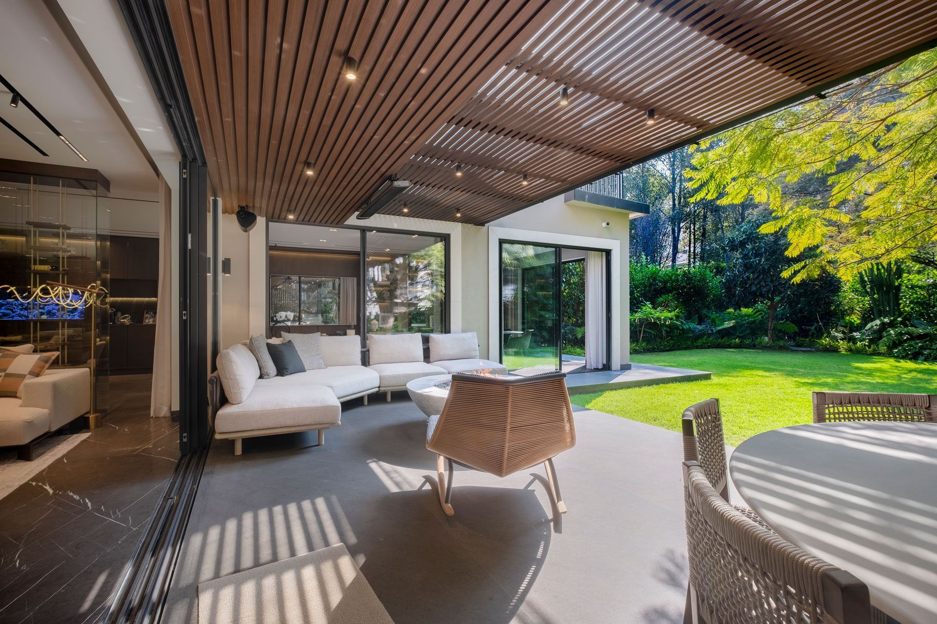 Patio moderno con sofás, techo con listones de madera y vistas a un jardín verde a través de puertas de cristal abiertas.