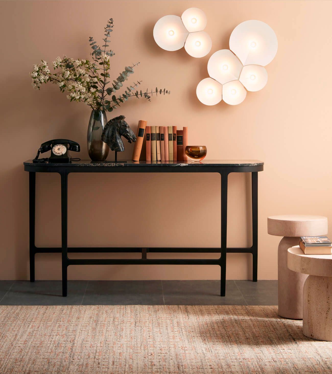 Sala de estar minimalista con mesa consola negra, libros, jarrón, apliques de pared y alfombra tejida sobre una pared beige.