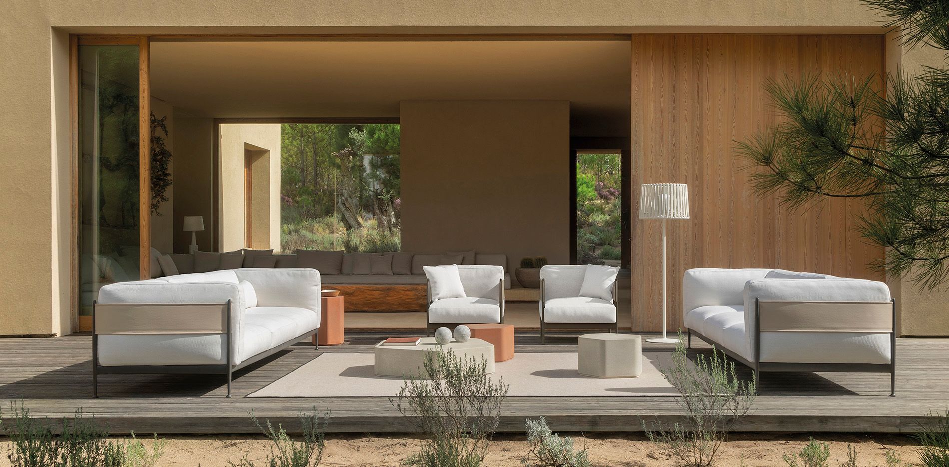 Patio moderno con asientos blancos, mesas bajas y un salón de estuco beige con vistas al jardín.