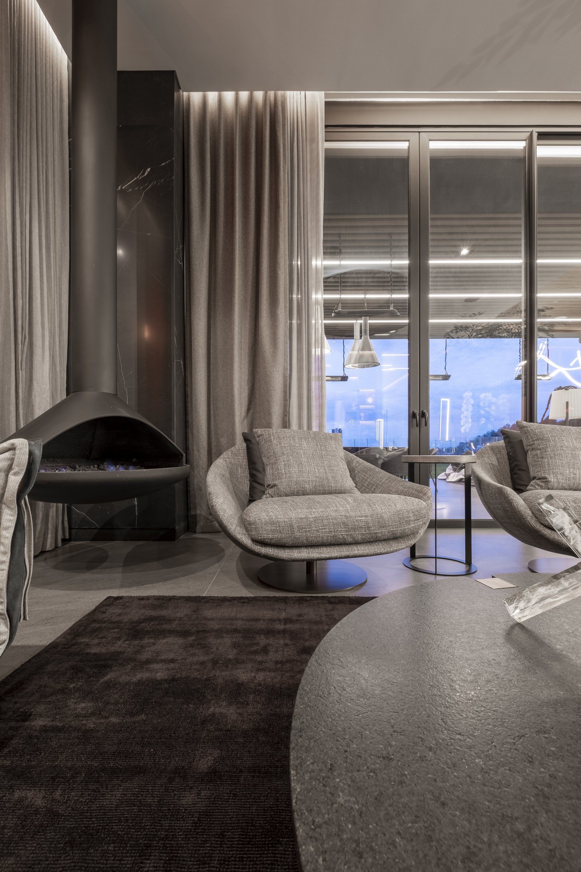 Salón moderno con sillas curvas, chimenea oscura y ventanales del suelo al techo con vistas a un paisaje nevado.