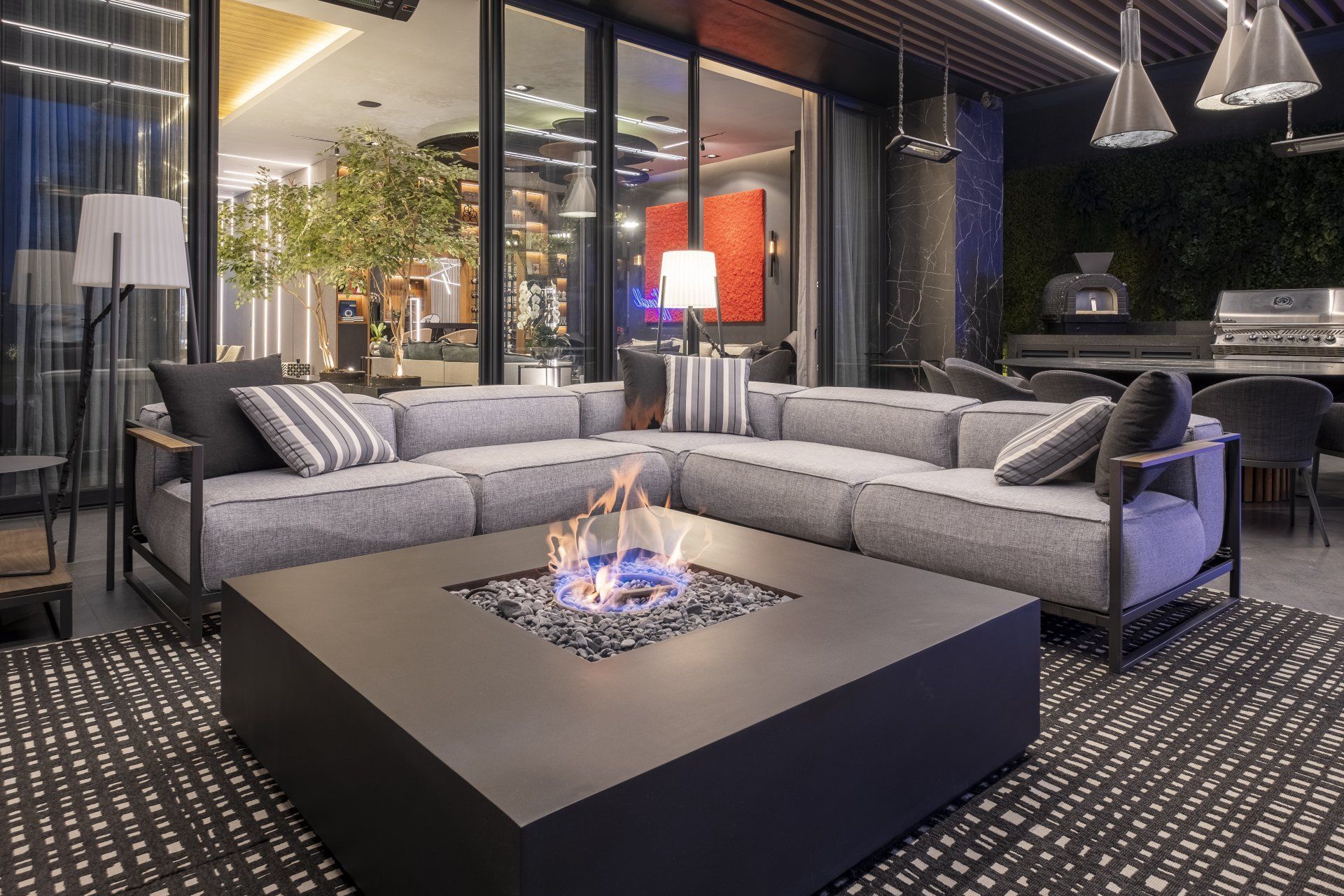 Terraza moderna en la azotea con sofás seccionales grises alrededor de una hoguera cuadrada bajo luces colgantes.