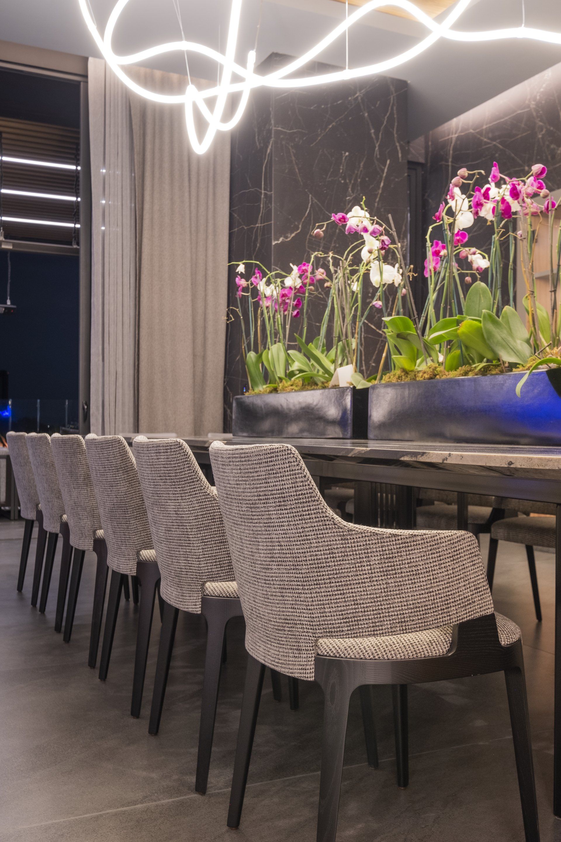 Comedor de restaurante moderno con sillas de mimbre, mesa larga y orquídeas rosas bajo una luz de techo brillante.