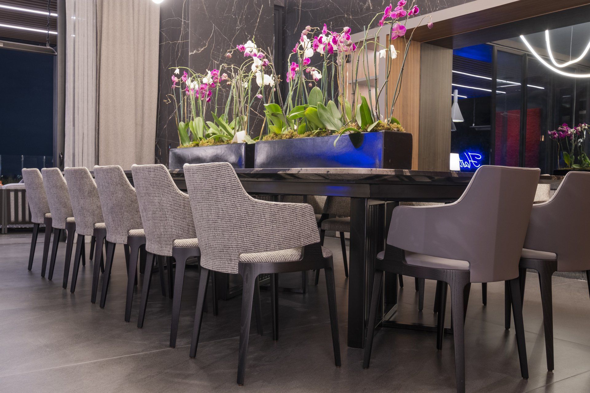 Una hilera de sillas de mimbre rodea una mesa alta con orquídeas en el salón de un restaurante moderno.