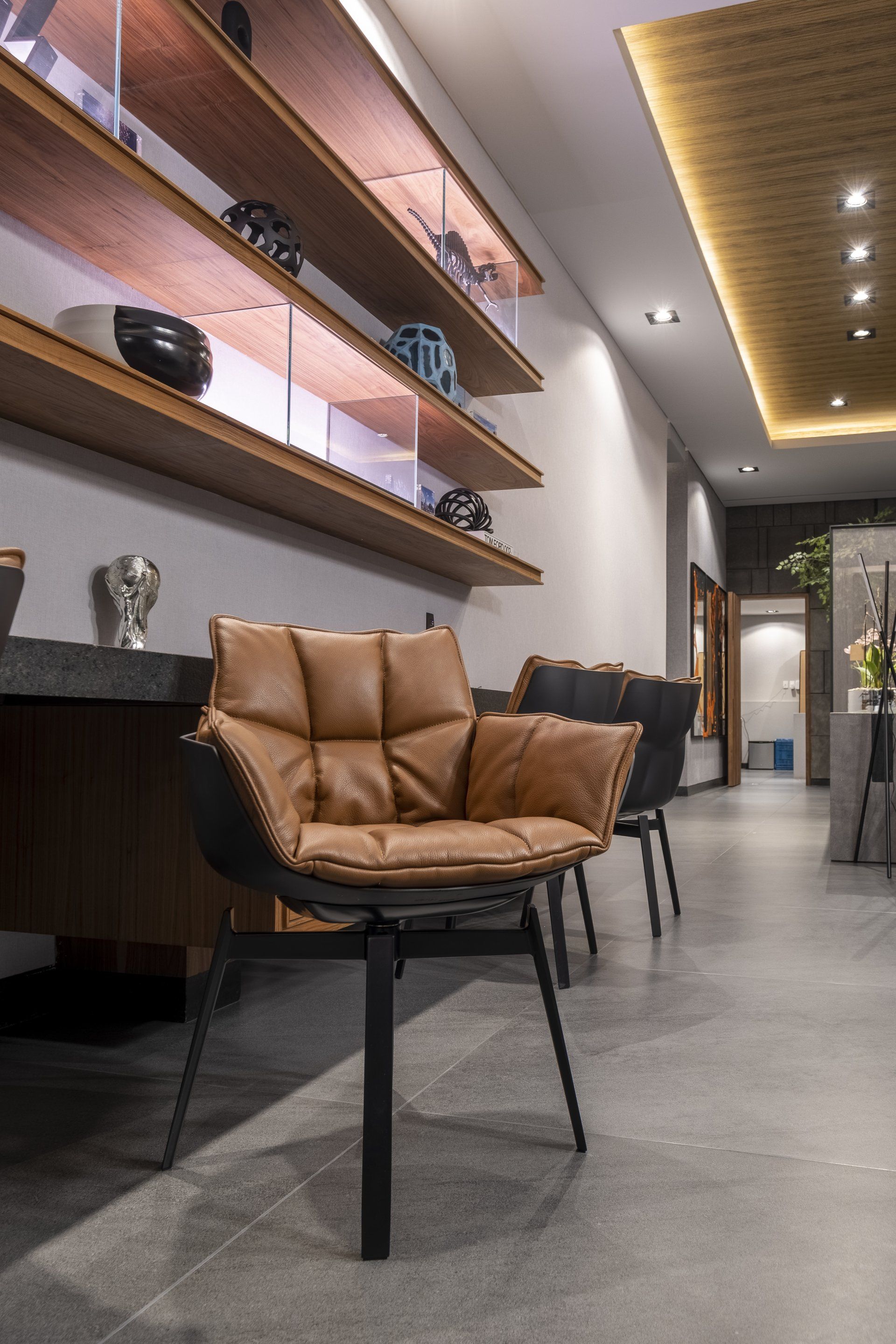 Pasillo moderno con estanterías, silla color canela, suelo gris y luces de techo cálidas.