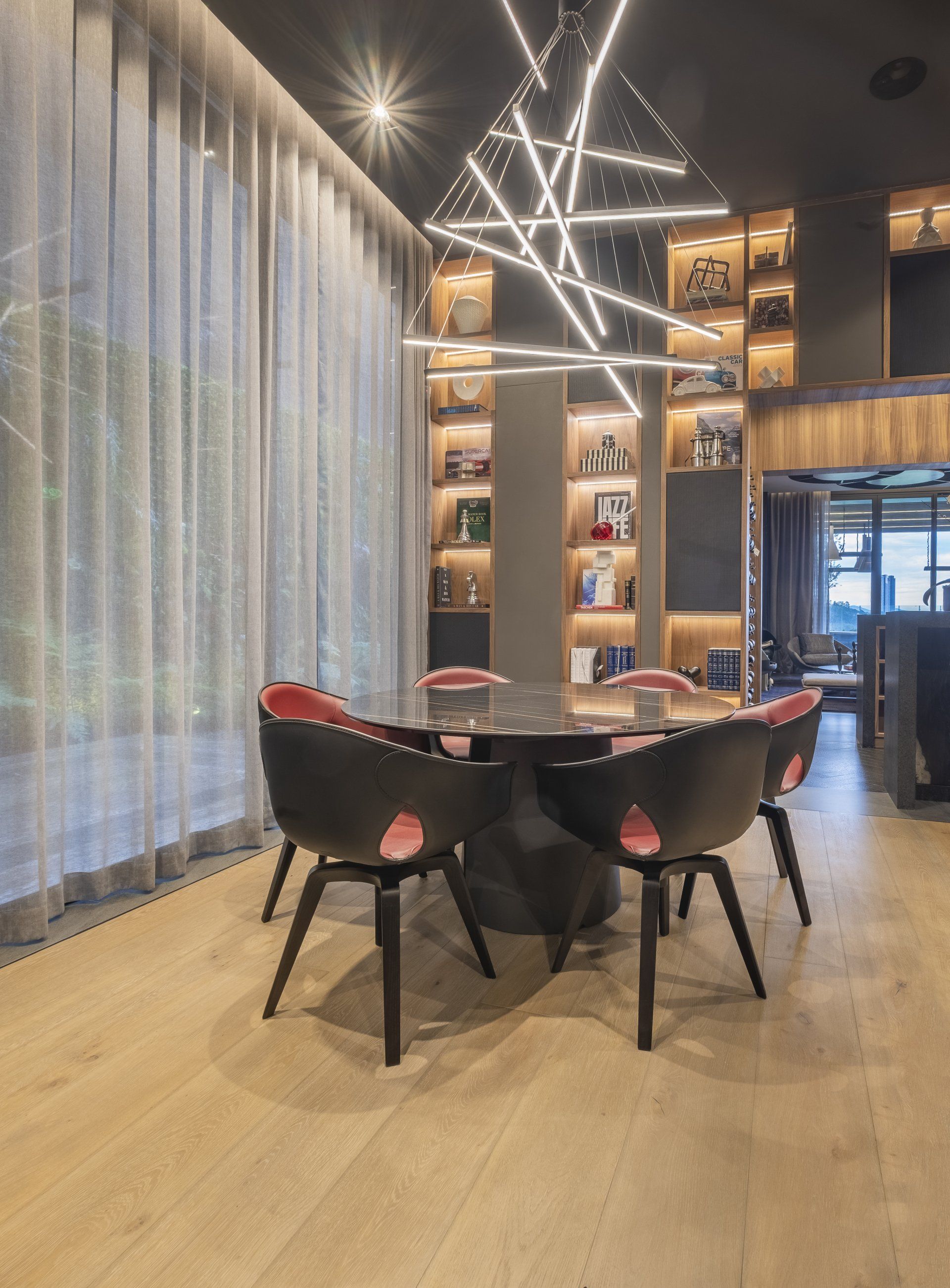 Salón moderno con sillas negras, mesa redonda, estantes de madera, lámparas colgantes y cortinas altas transparentes.