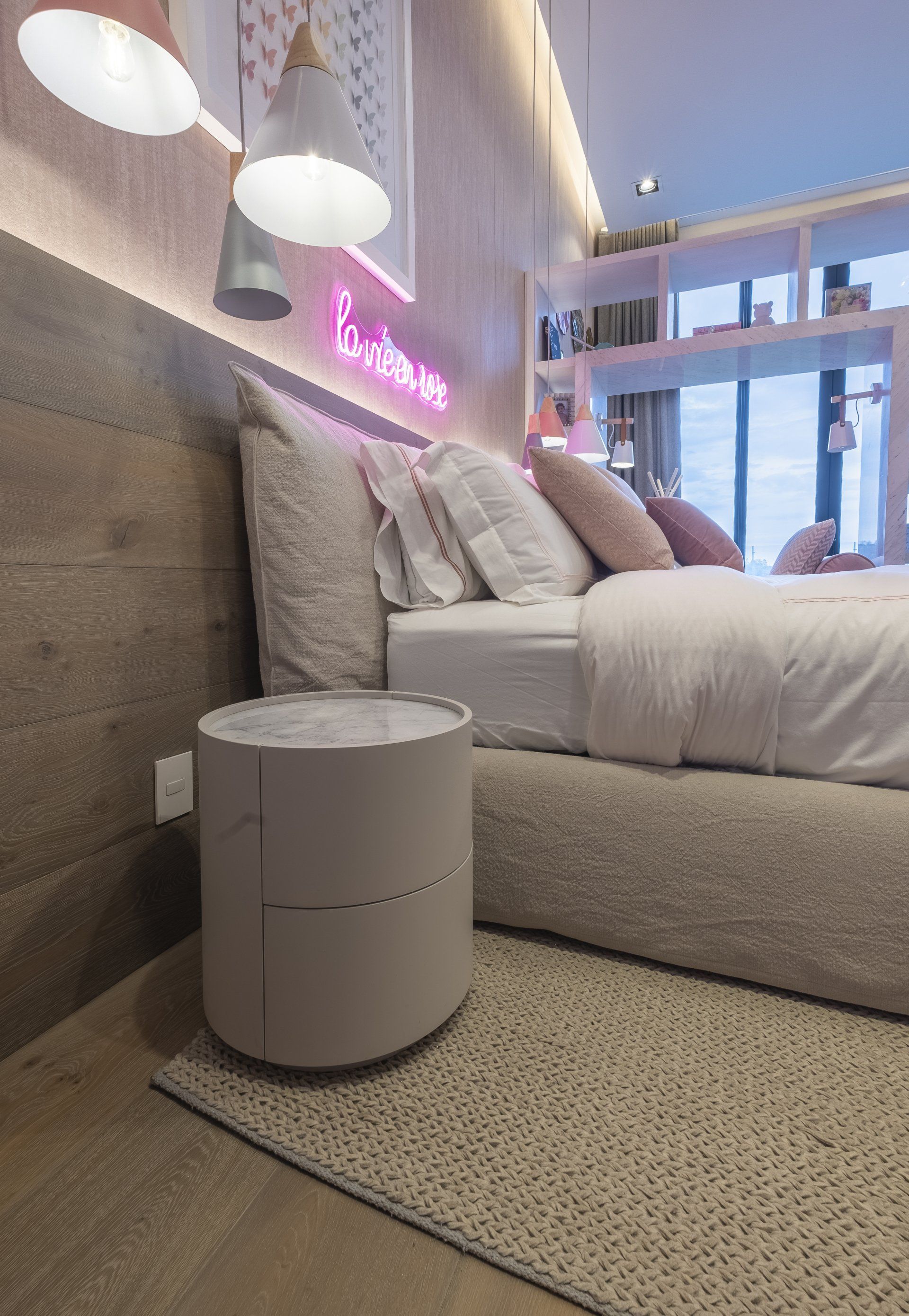 Habitación acogedora con cama, lámparas, mesita de noche redonda gris y letrero de neón rosa en la pared.