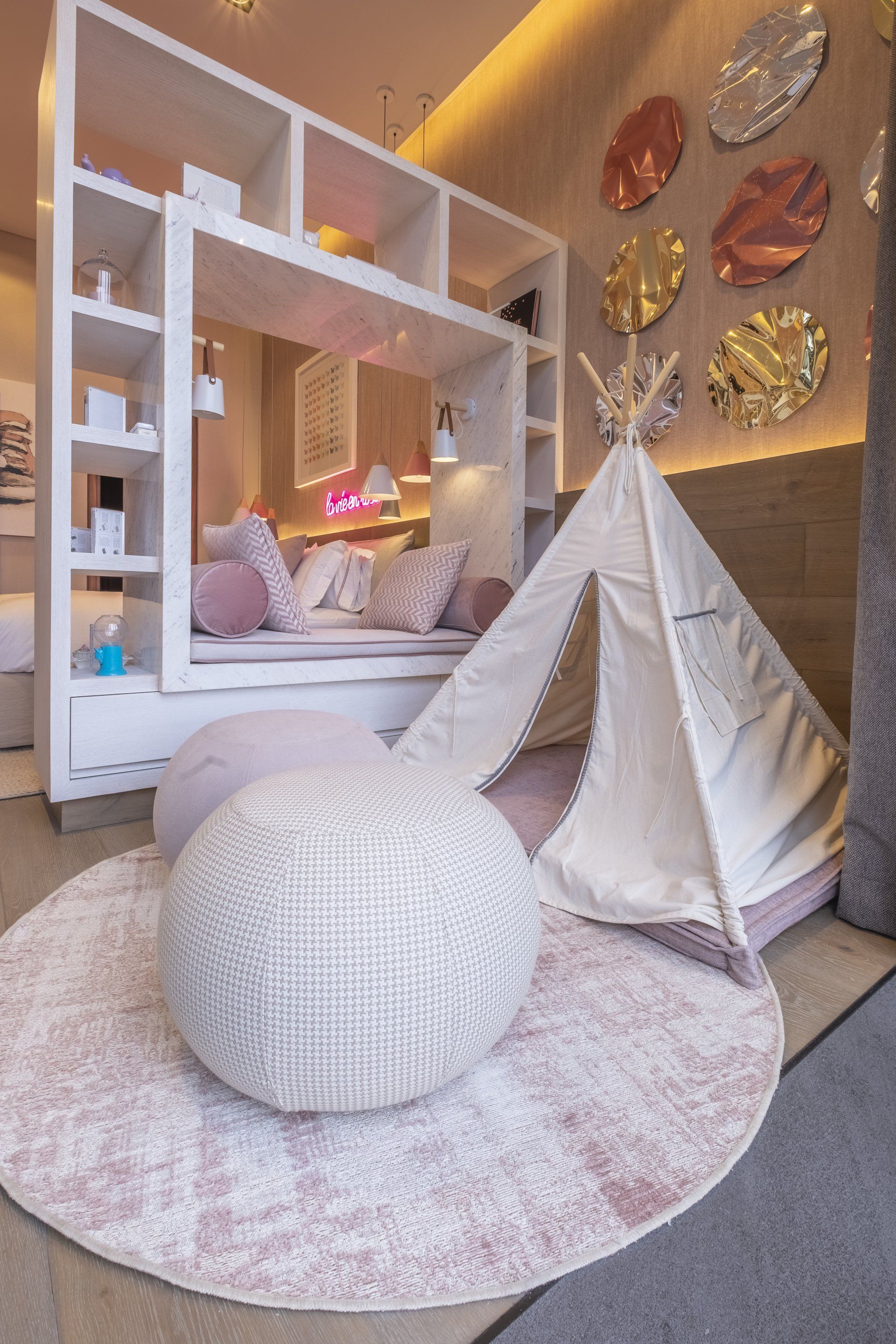 Rincón de juegos para niños con un tipi blanco, un cojín redondo y una alfombra rosa bajo una cálida decoración de pared.