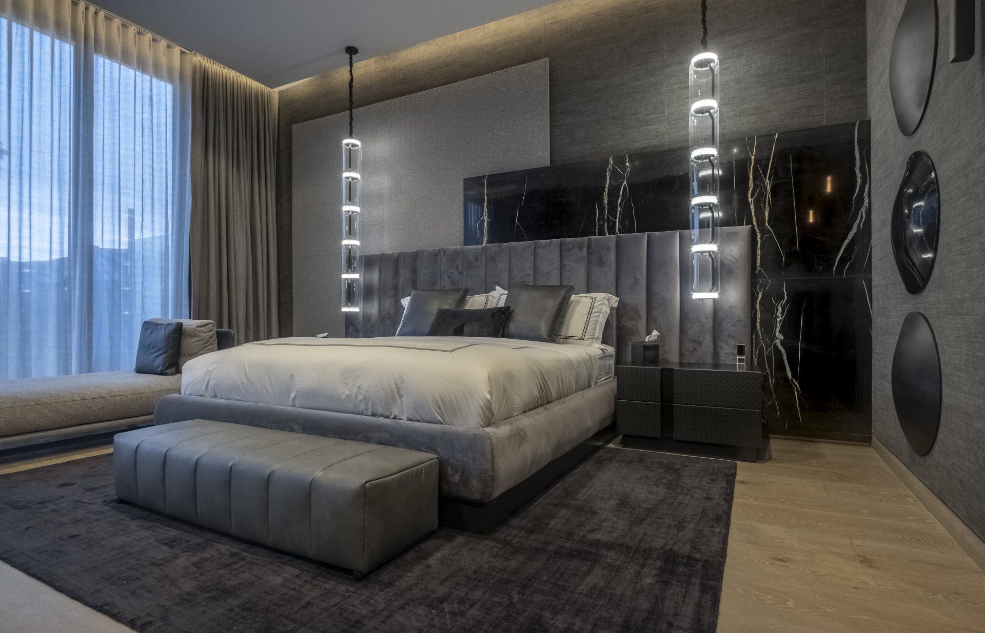 Dormitorio moderno en tonos grises con una cama grande, banco capitoné, ventanales del suelo al techo y lámparas colgantes.