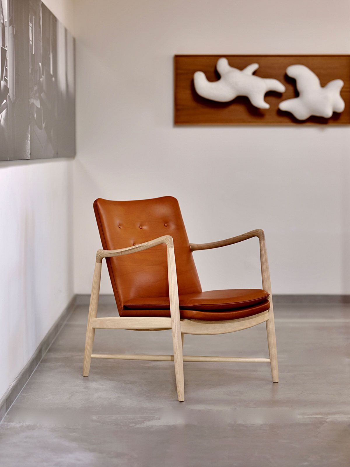 Sillón auxiliar de cuero color canela en una habitación minimalista con paredes blancas y suelo gris, junto a un cuadro.