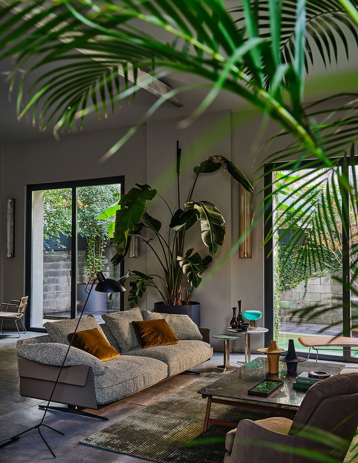 Sala de estar moderna con sofá gris, cojines estampados, plantas y grandes ventanales con vistas a un patio tropical.