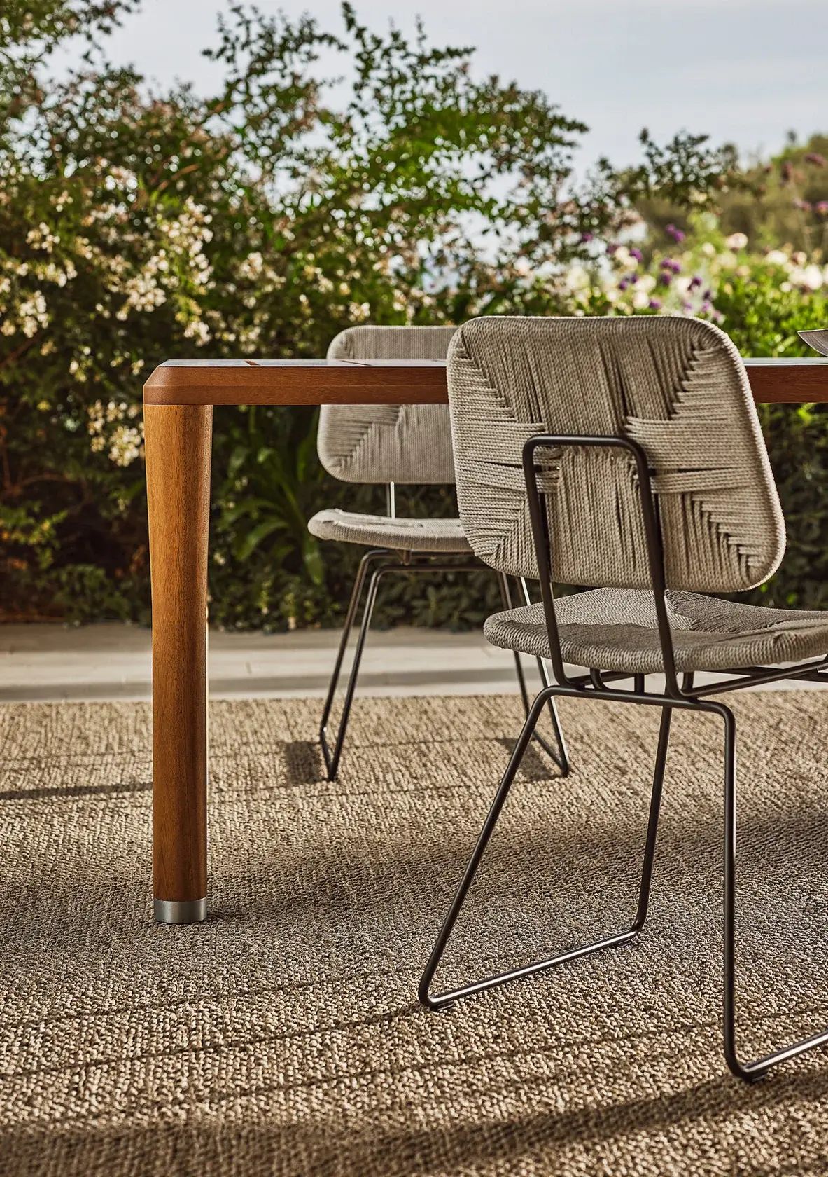 Dos sillas de mimbre junto a una mesa de madera en un patio soleado con árboles al fondo.