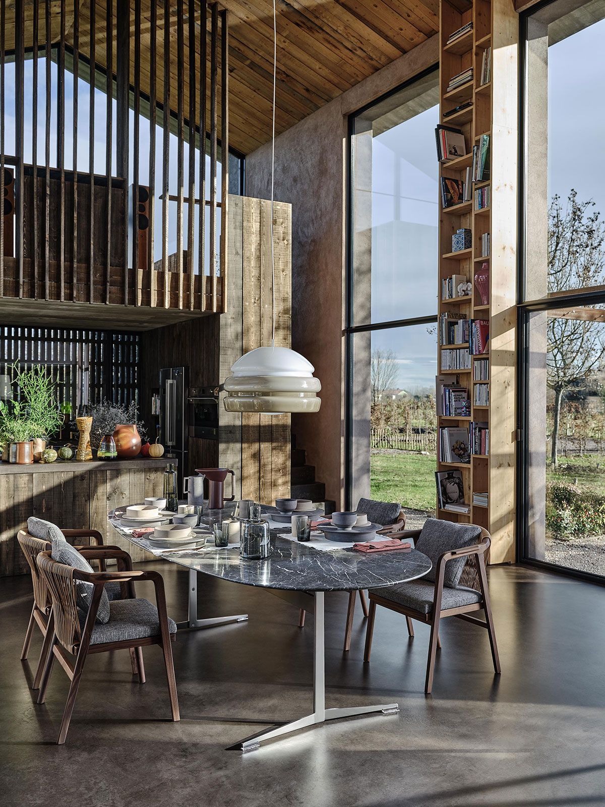 Comedor moderno con mesa redonda, sillas de mimbre y grandes ventanales que van del suelo al techo con vistas a la montaña.