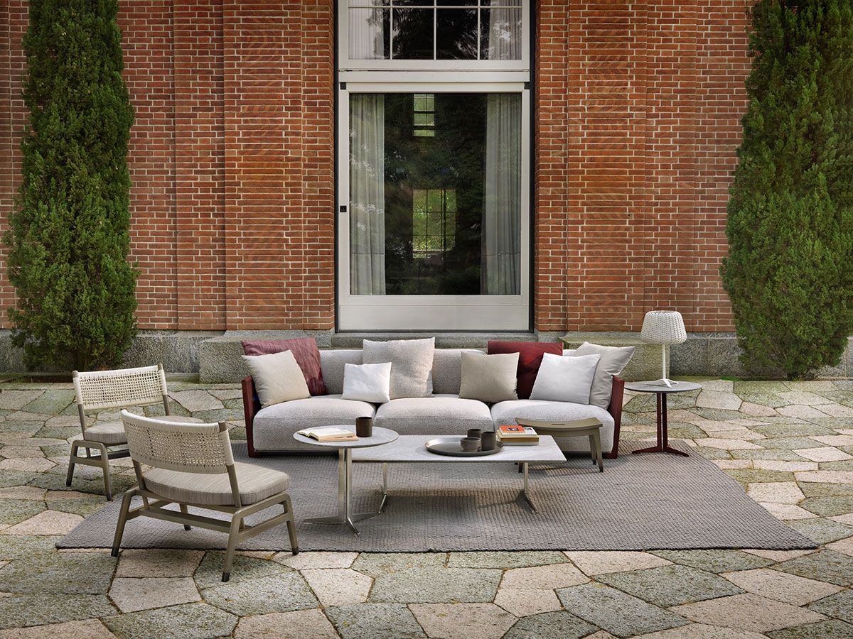 Patio exterior con sofá gris, sillas y mesa de centro sobre una alfombra, frente a una pared de ladrillo y una ventana.