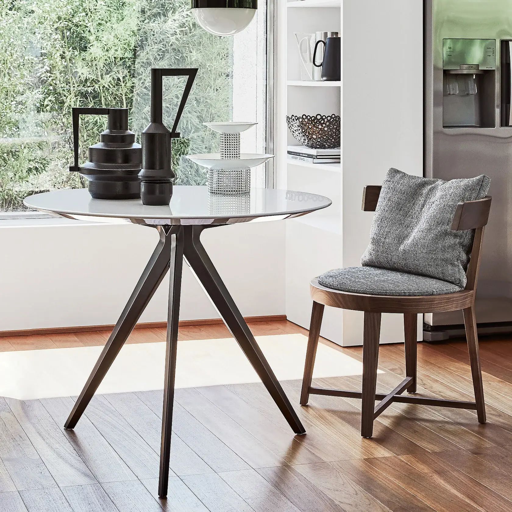 Rincón de comedor moderno con una mesa redonda de cristal, sillas negras y una ventana que deja entrar la luz del sol.