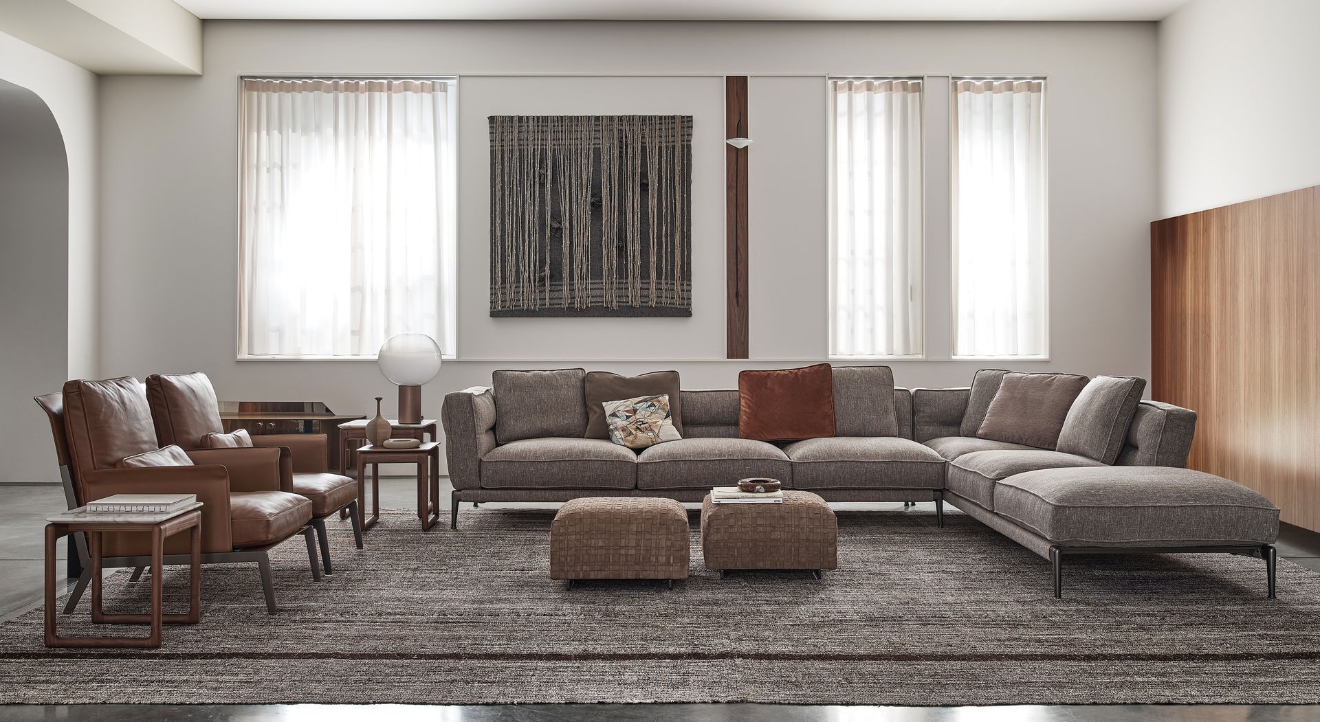 Sala de estar moderna con sofá seccional gris, alfombra estampada, sillas y grandes ventanales con cortinas transparentes.