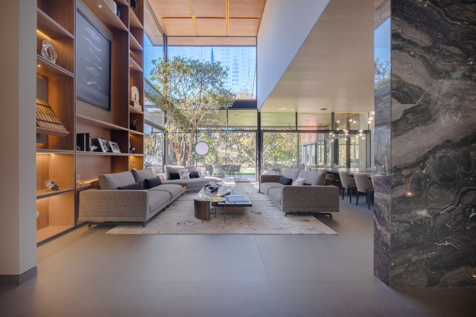 Salón moderno con sofás grises, una mesa de centro y ventanales que van del suelo al techo con vistas a los árboles.