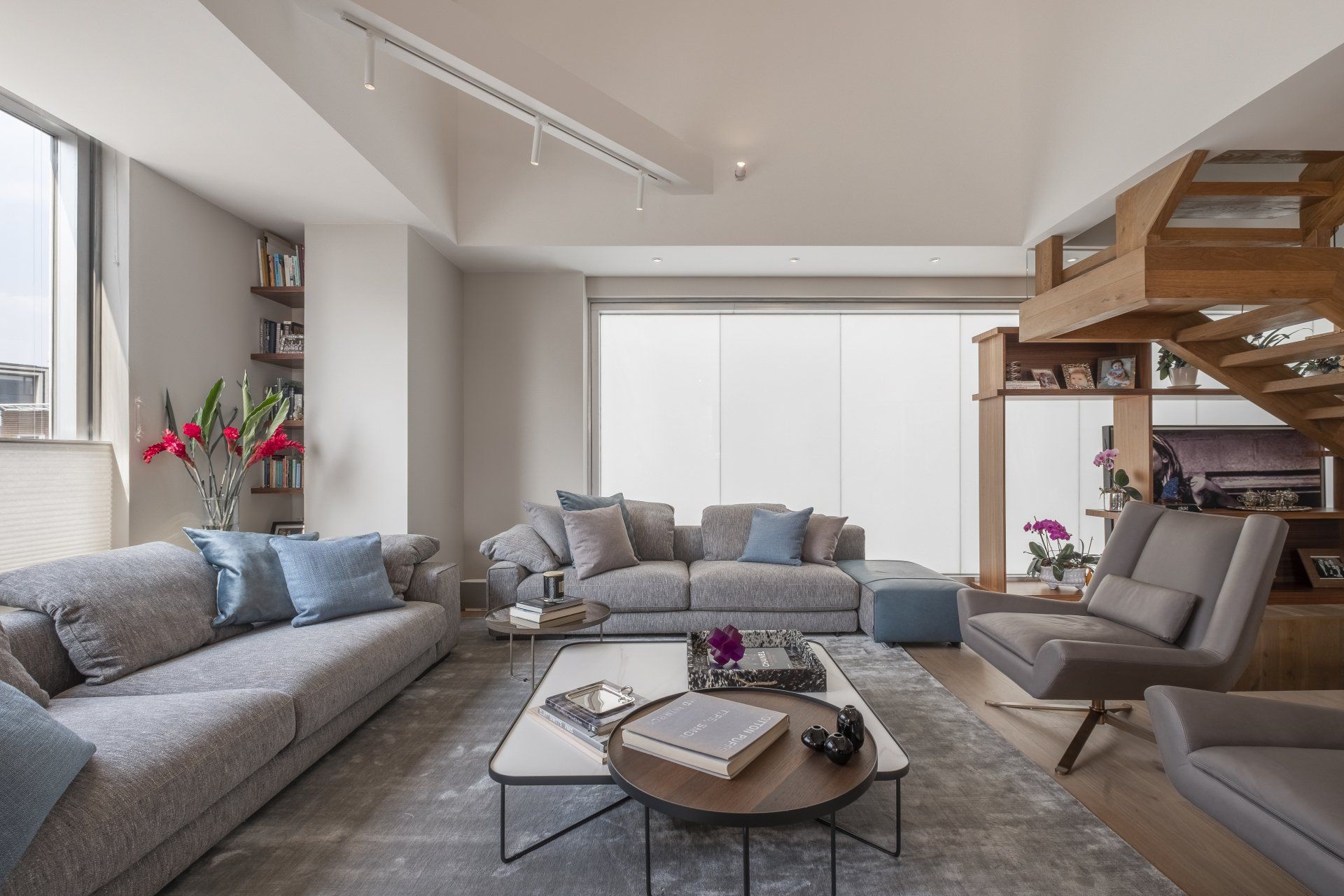Sala de estar moderna con sofás grises, una mesa de centro, lámpara de pie y escalera de madera junto a grandes ventanales.