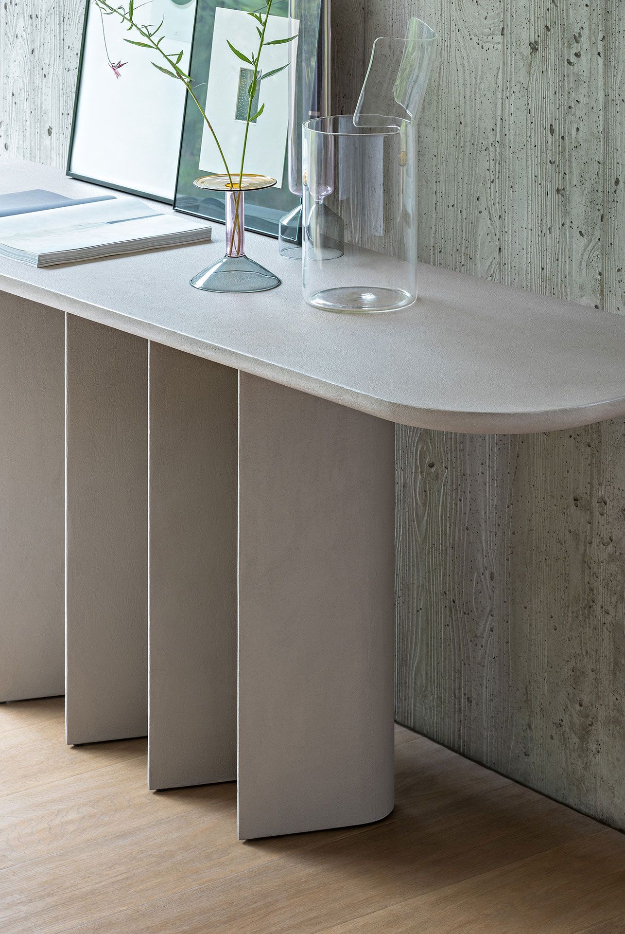 Mesa consola moderna con bordes curvos, patas de tres paneles y jarrones contra una pared texturizada.