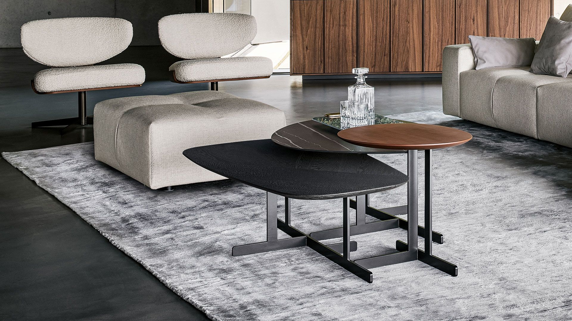 Sala de estar moderna con sofás grises, sillones beige y mesas de centro negras y de madera apiladas sobre una alfombra