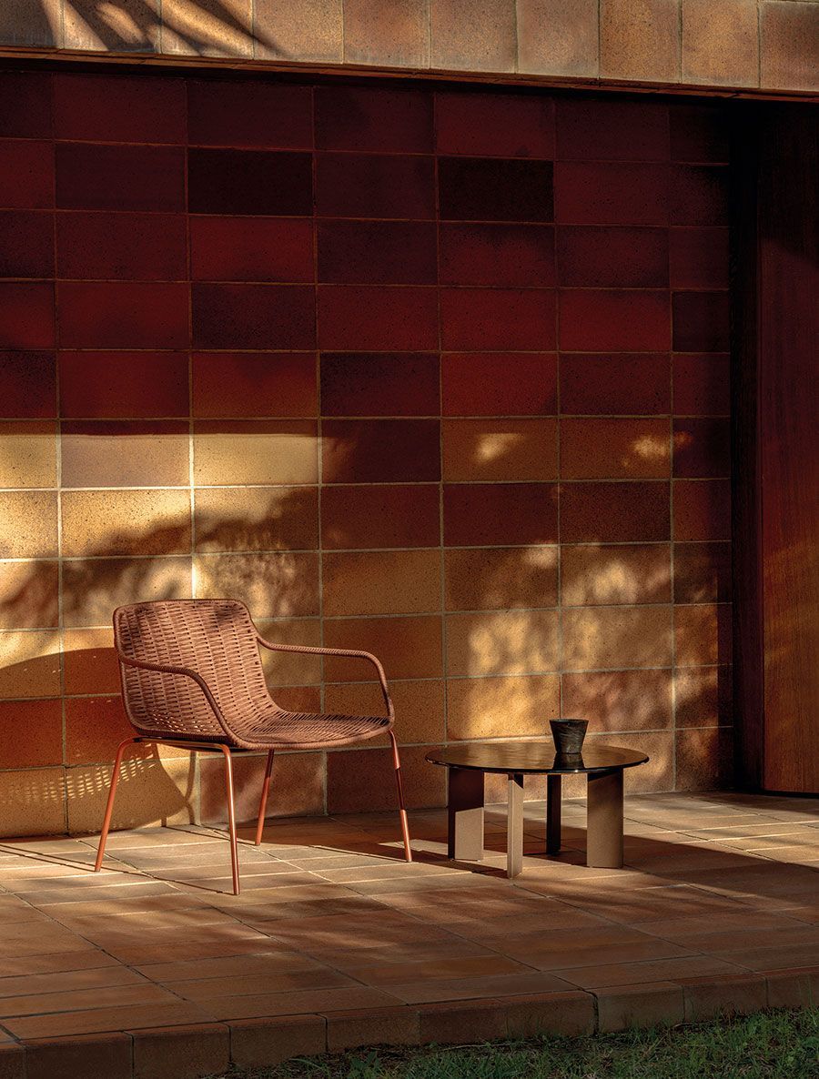 Silla de mimbre y mesita en un patio de madera, frente a una pared de azulejos rojos iluminada por el sol y con sombras.