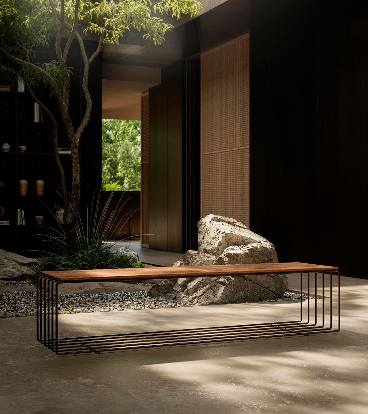 Patio moderno con un largo banco de madera, base tejida, pieza central de piedra y luz solar tamizada bajo los árboles.